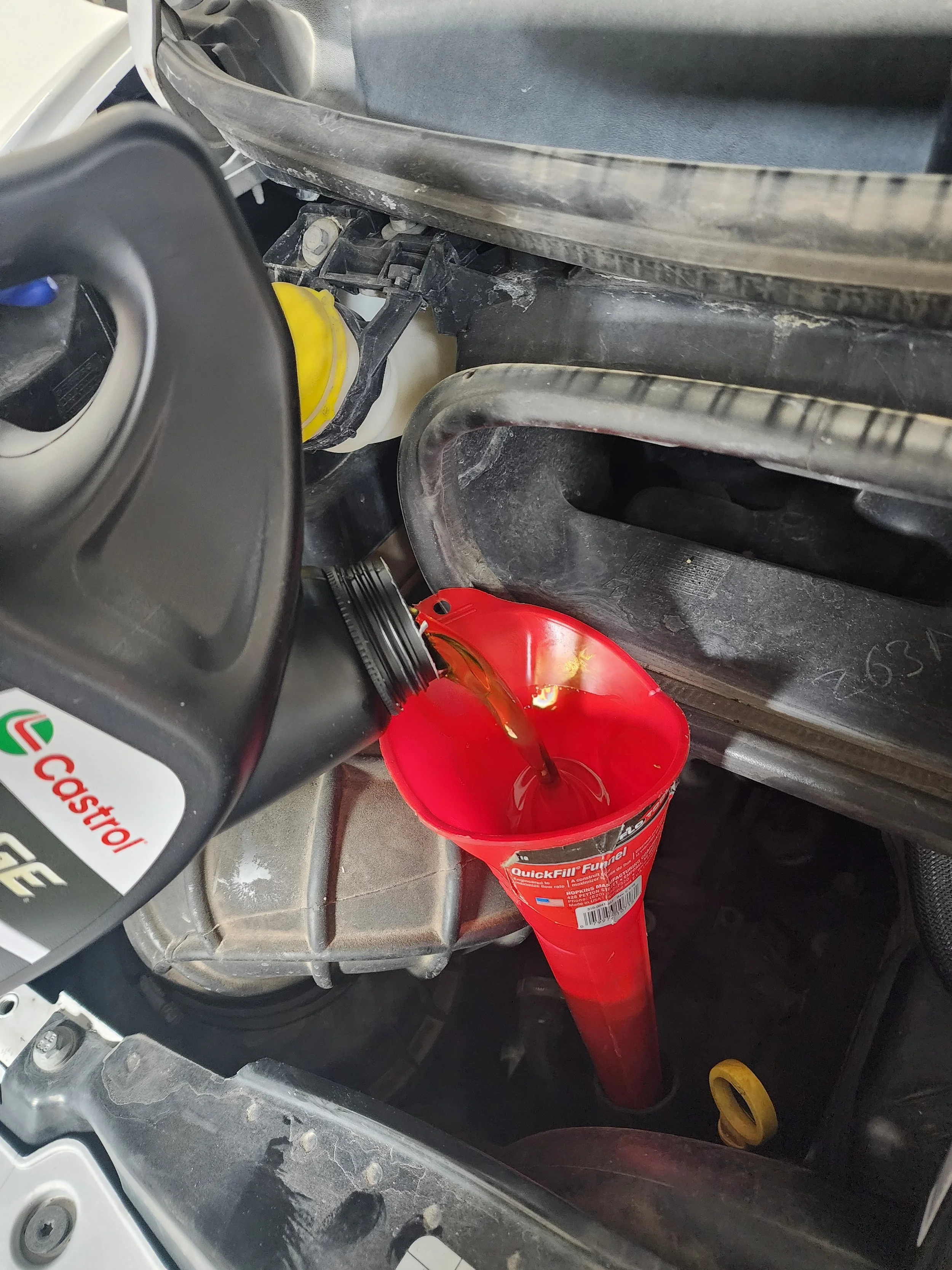 A red funnel positioned under a bottle of Castrol motor oil, with oil pouring from the bottle into the funnel in a car's engine compartment.