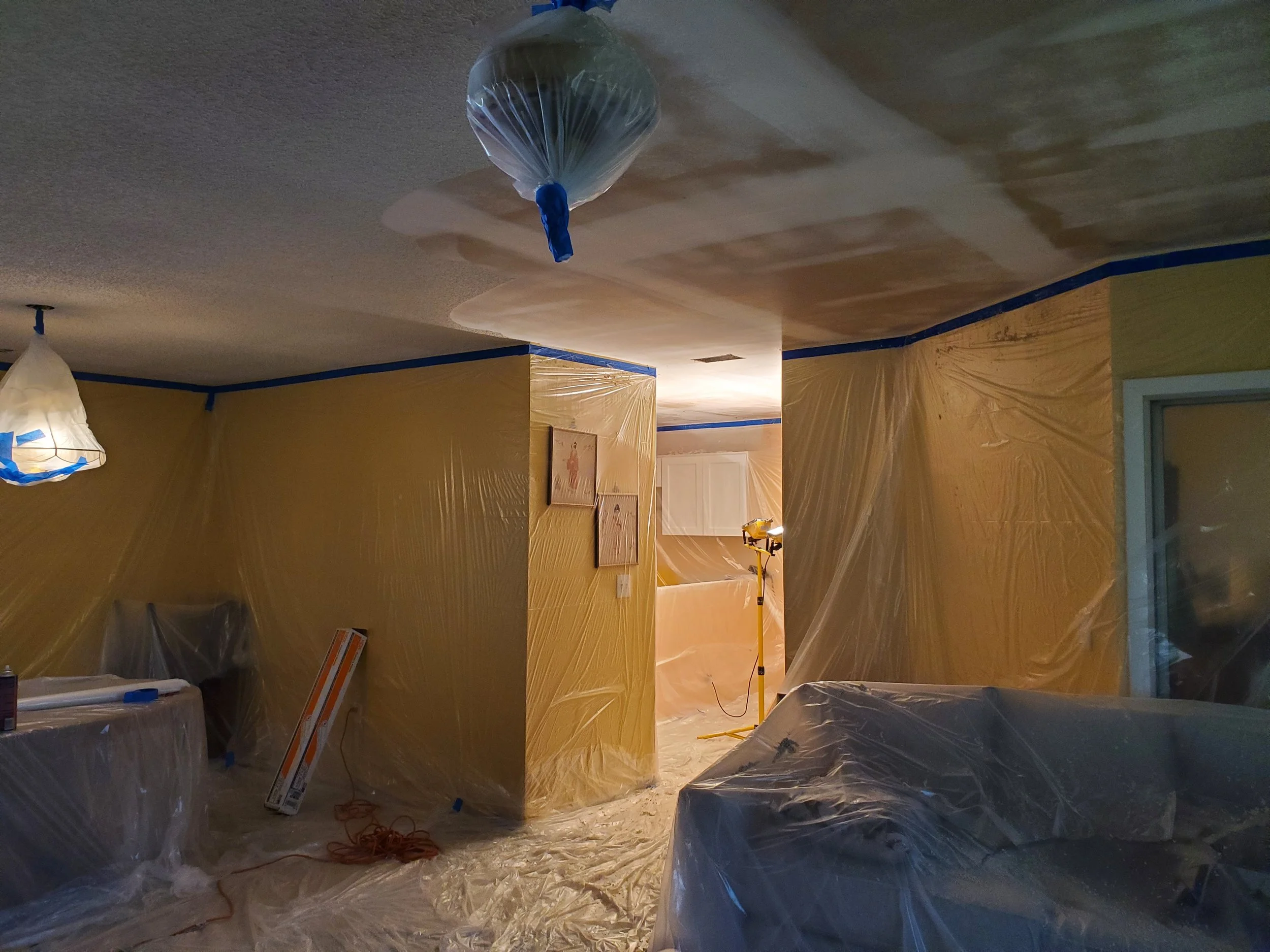 Living room during a popcorn ceiling removal, with plastic sheeting covering furniture, walls, and floors. A temporary work light to ensure quality before knockdown texture is applied and ceiling is painted.