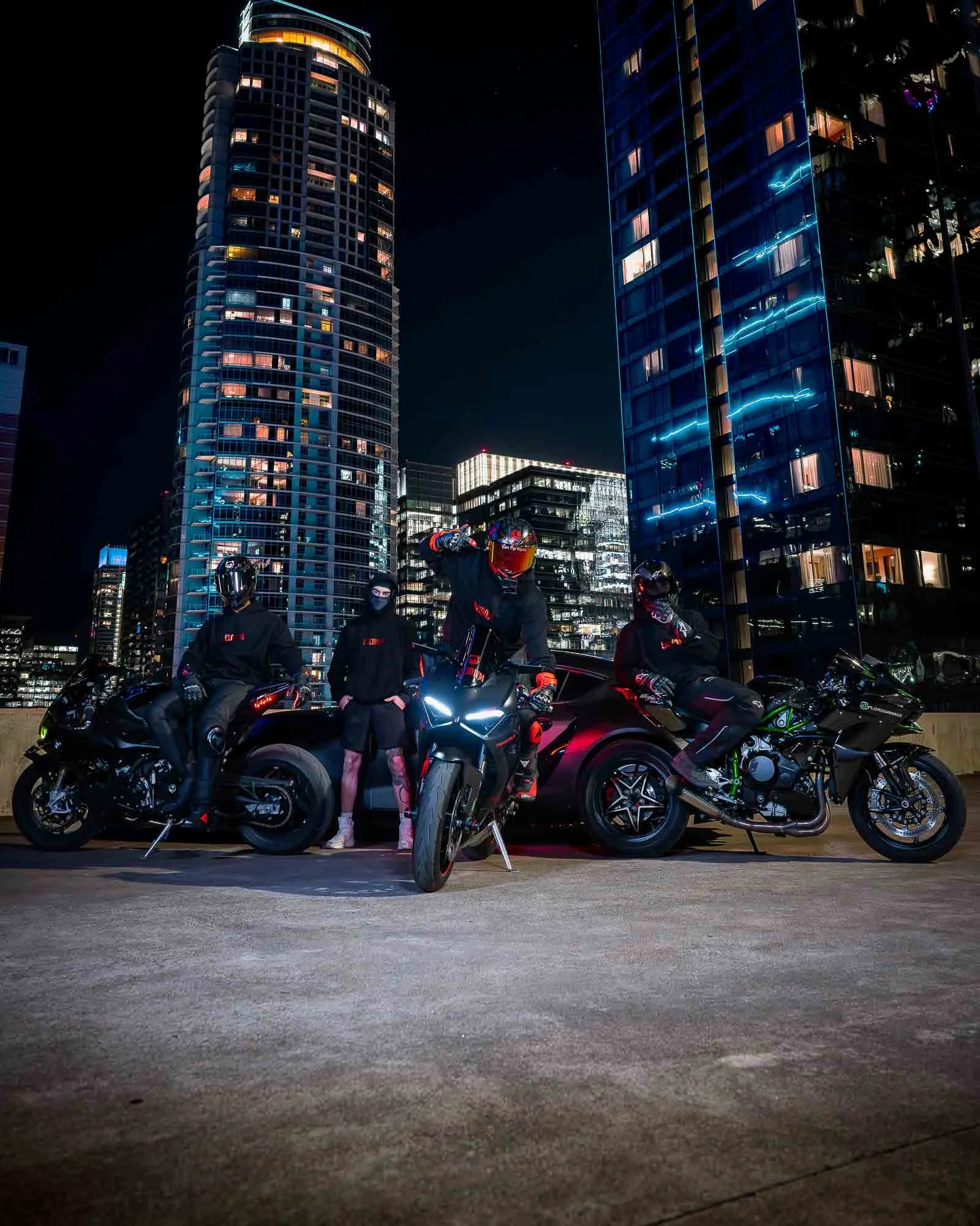 EXCIDIVM riders with sport motorcycles wearing black streetwear in city skyline at night