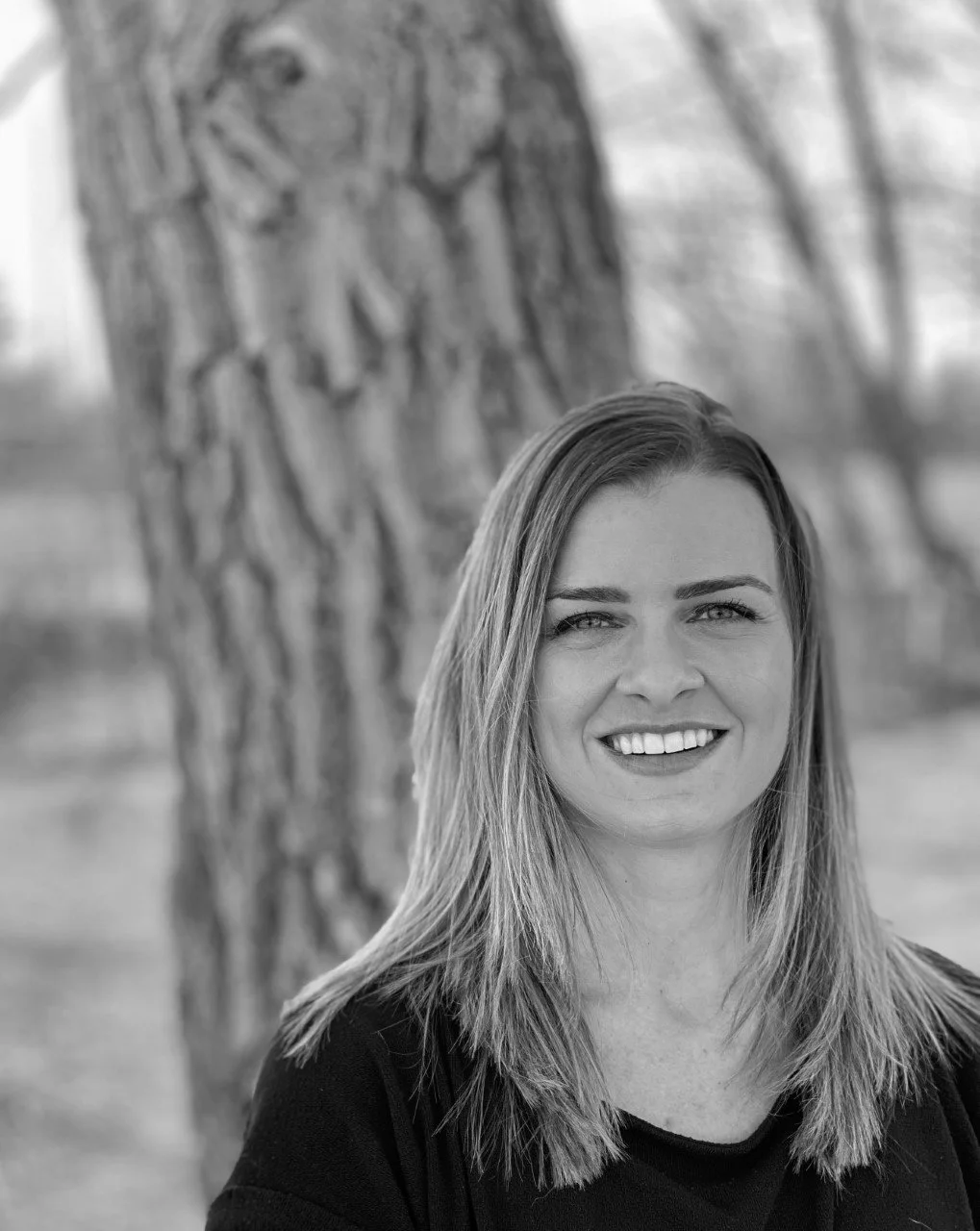 A smiling woman with straight hair standing outdoors near a tree trunk.