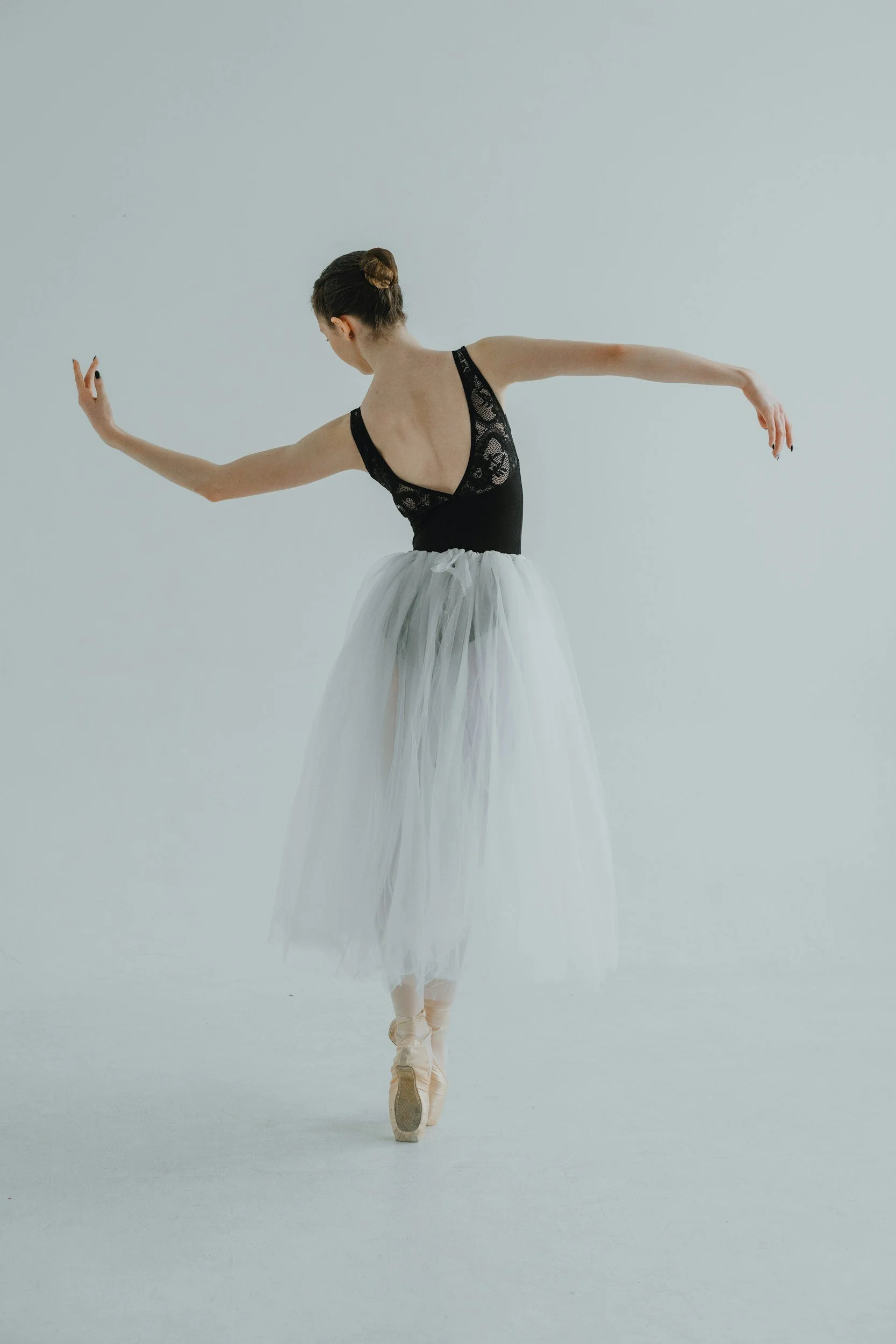 A ballerina in a black lace leotard and white tutu practicing ballet on pointe, performing a graceful pose against a plain light background.