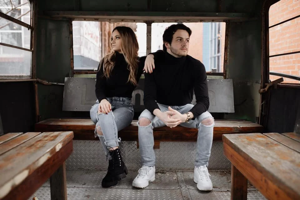 A young man and woman sitting on a bench inside a rustic bus with wooden seats, metal flooring, and windows showing city buildings outside.