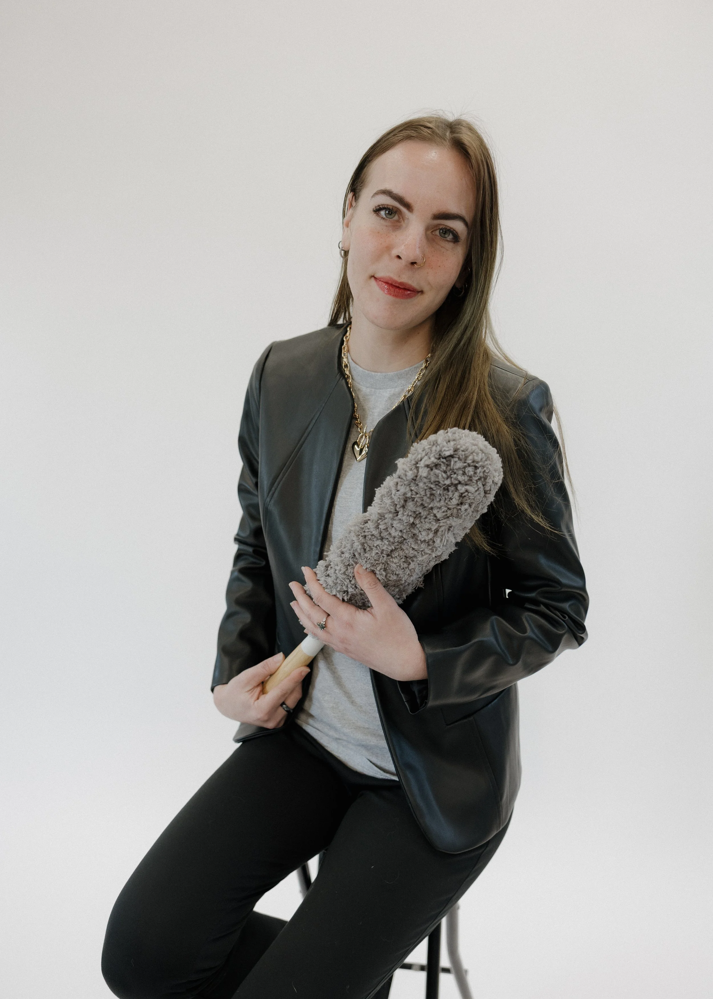 A young woman with long brown hair, wearing a black leather jacket, light gray t-shirt, and black pants, sits on a stool against a plain white background. She is holding a gray duster with a wooden handle and has a slight smile on her face.