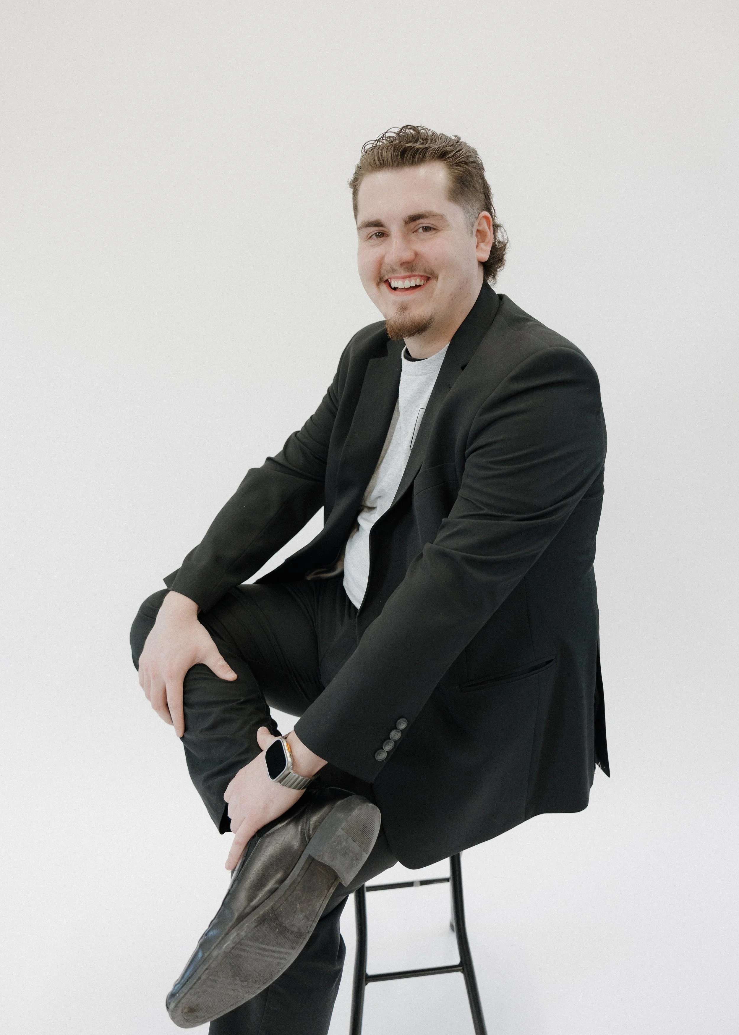 A young man with brown hair and a beard, dressed in a black blazer, white shirt, and black pants, sitting on a black stool against a plain white background, smiling and adjusting his shoe.