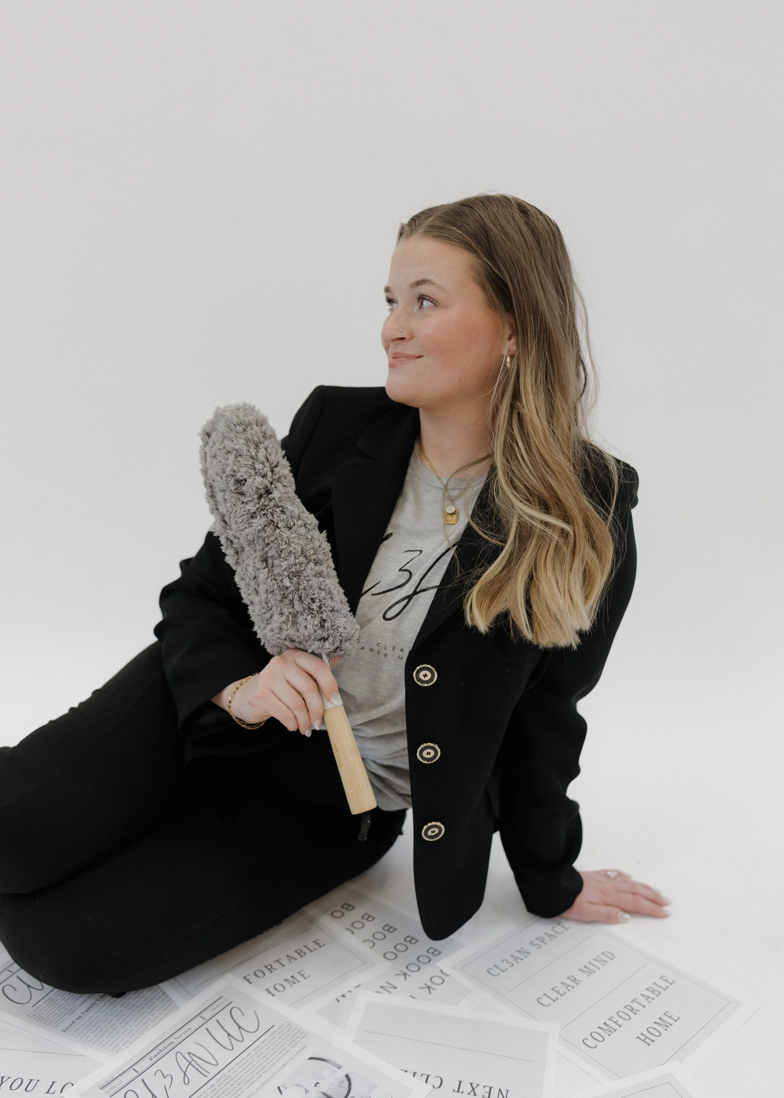 A young woman sitting on the floor, holding a duster, surrounded by papers with words like 'Comfortable Home' and 'Clear Mind', against a plain white background.