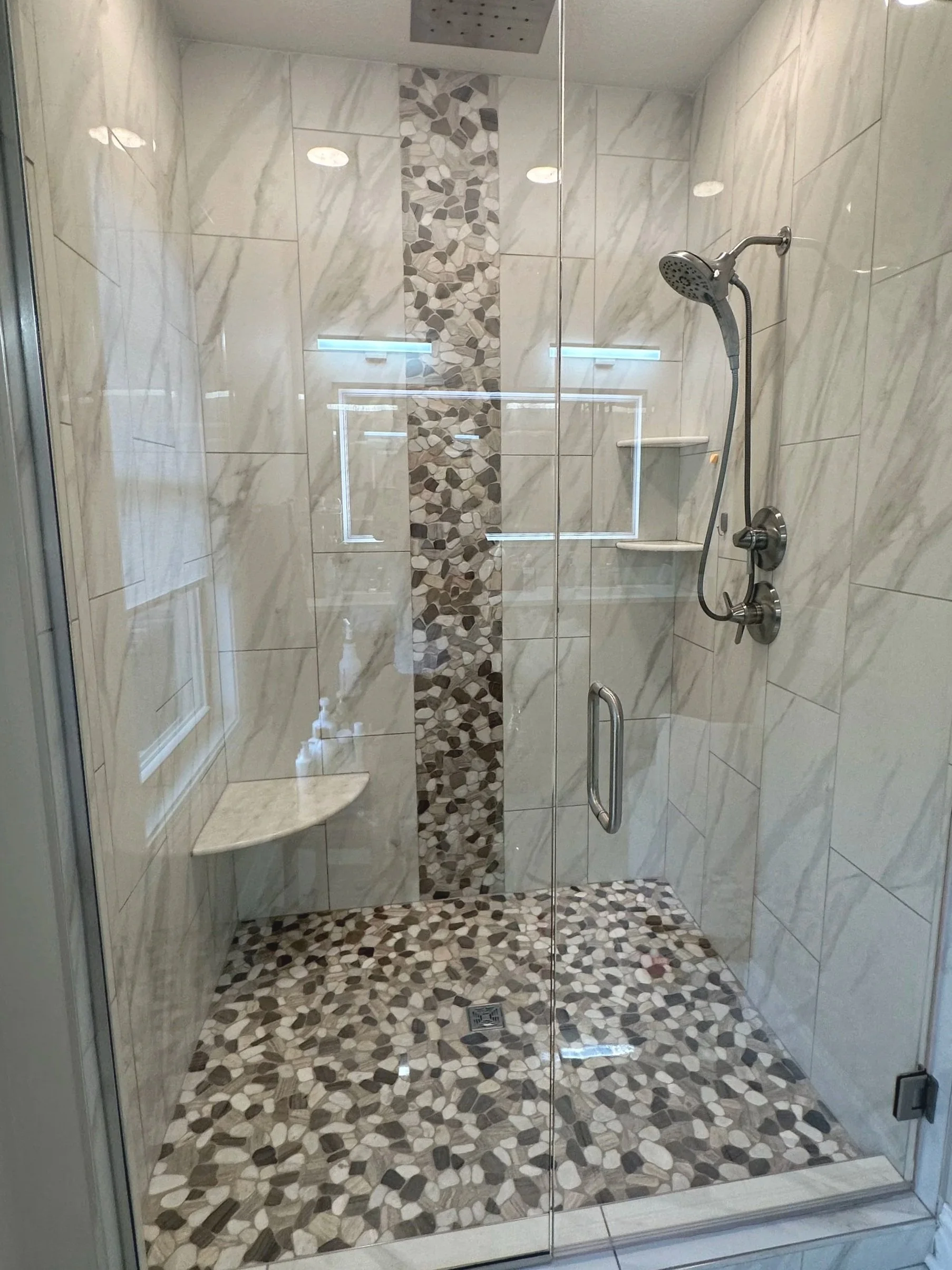 Glass shower enclosure with tiled walls and pebble-patterned floor and accent strip, two built-in corner shelves, and a chrome showerhead.