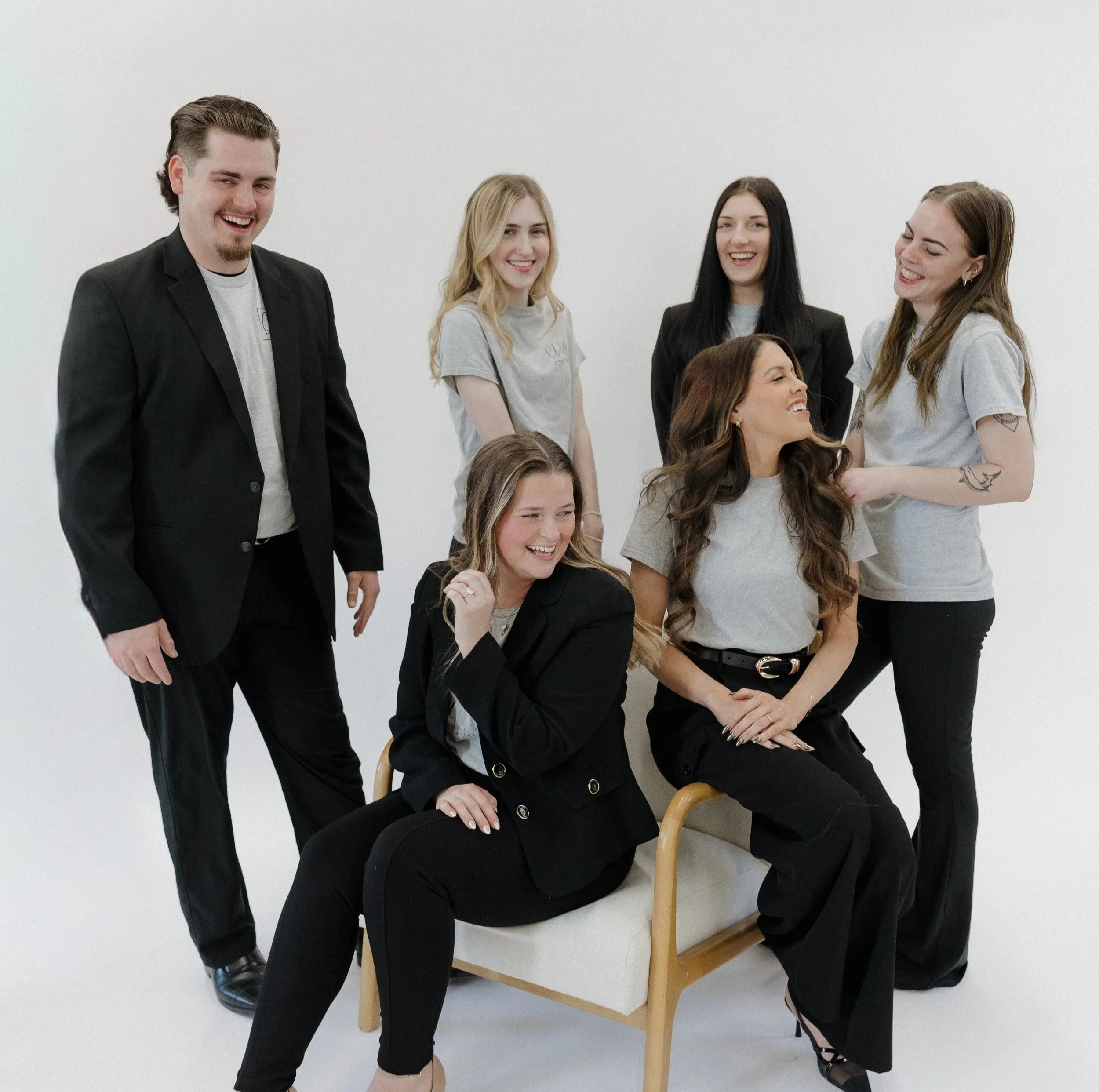Group of seven young adults, five women and two men, laughing and chatting against a plain white wall. Some are sitting, others standing.