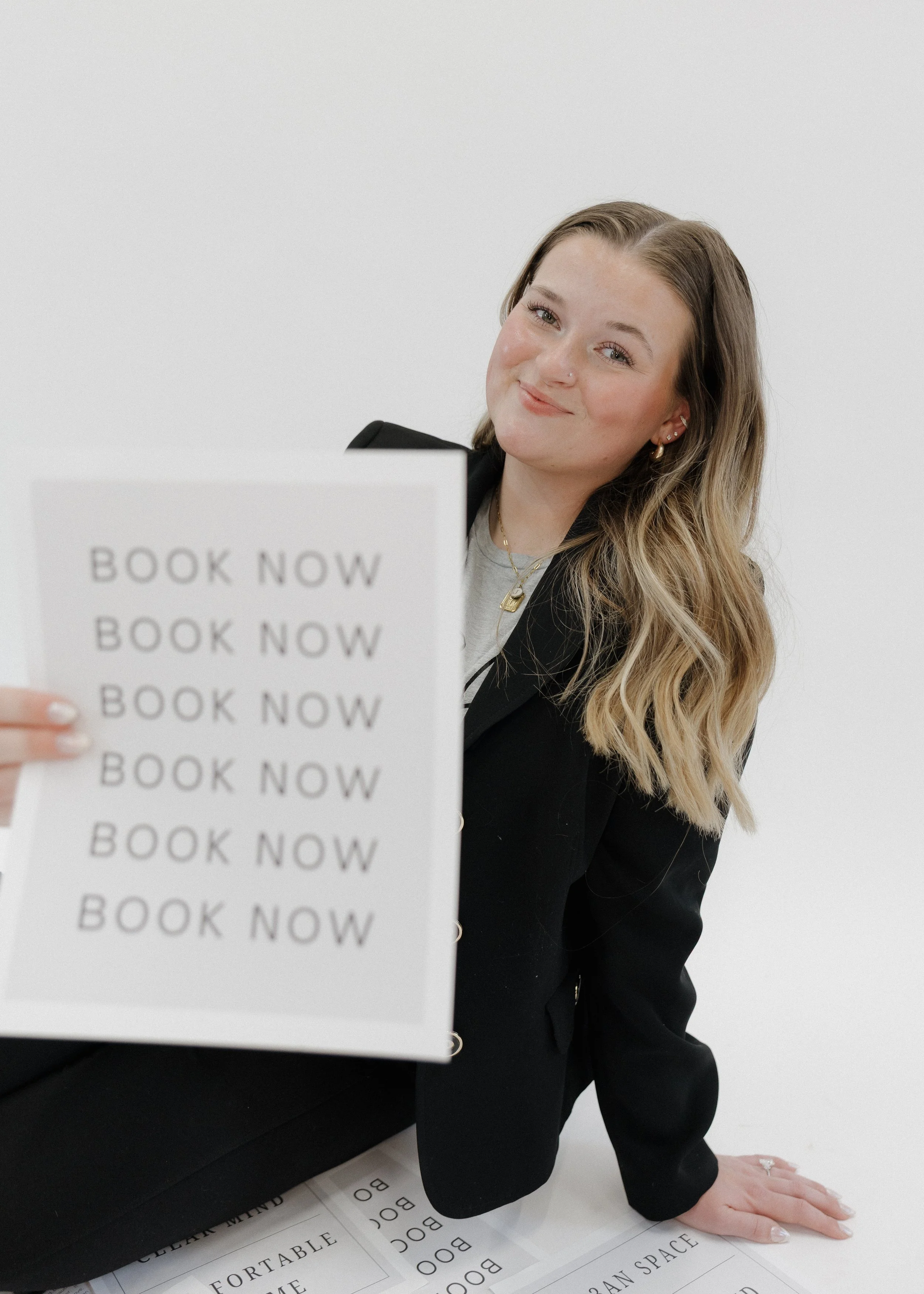 A smiling woman with wavy blonde hair holding a sign that says "BOOK NOW" multiple times, sitting against a plain white background.