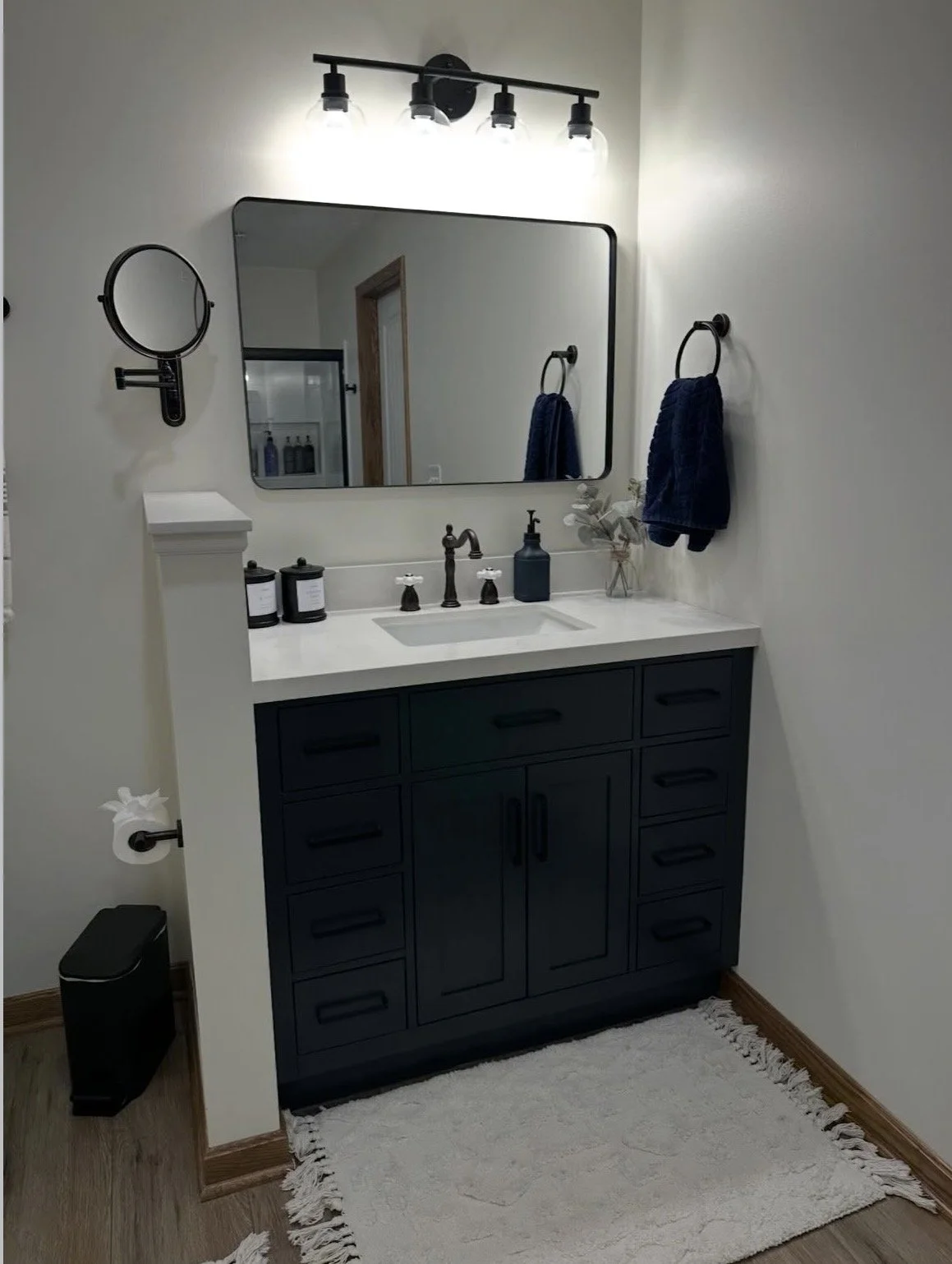 A modern bathroom vanity with a white countertop, dark blue cabinet, and a large mirror. There are black soap and lotion dispensers, a small vase with dried flowers, a wall-mounted magnifying mirror, two navy towels hanging on side rings, and four overhead light bulbs above the mirror. A white rug is on the wooden floor.