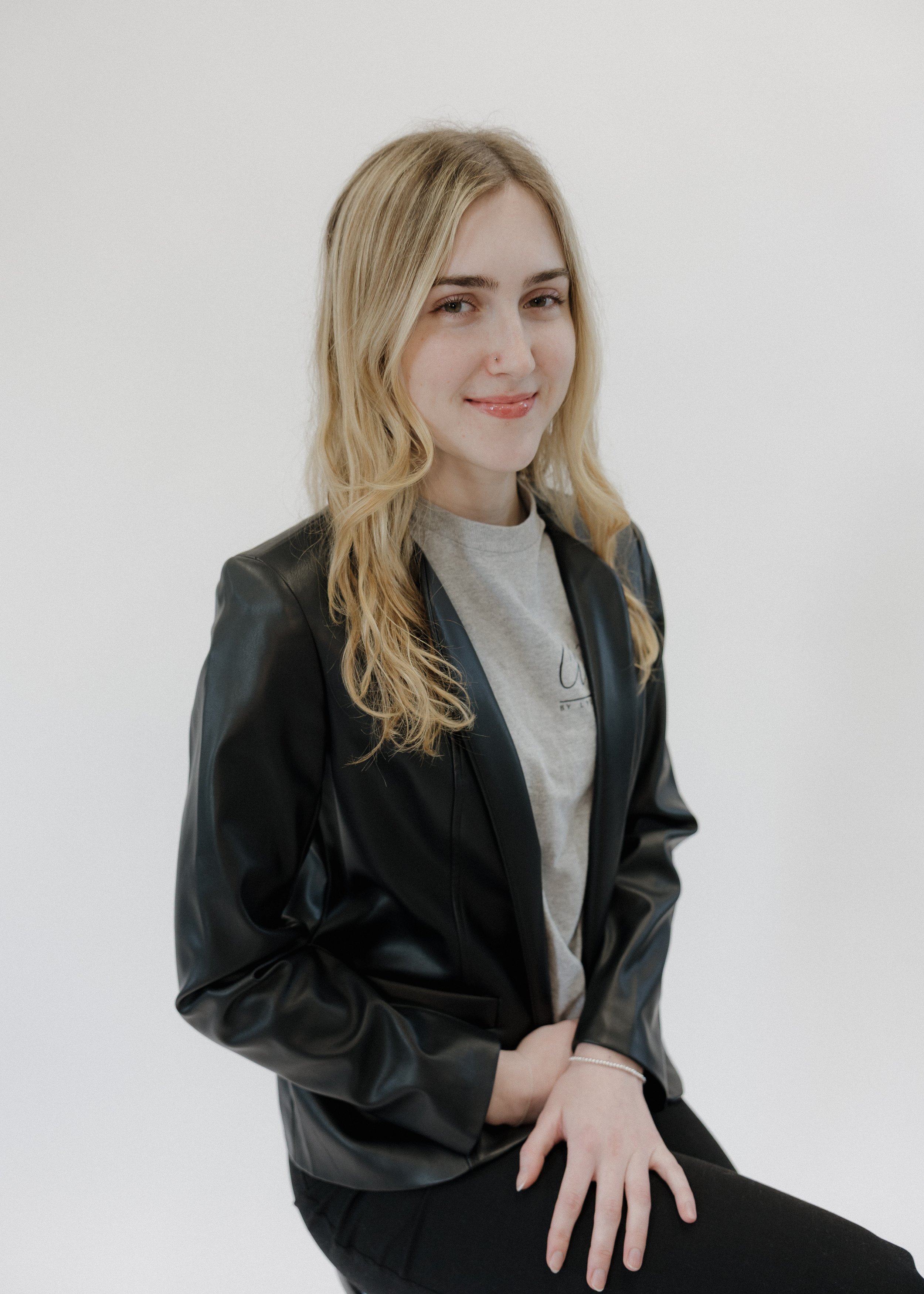 A young woman with blonde wavy hair and light skin, wearing a black leather jacket over a gray t-shirt, sitting against a plain white background, smiling softly at the camera.