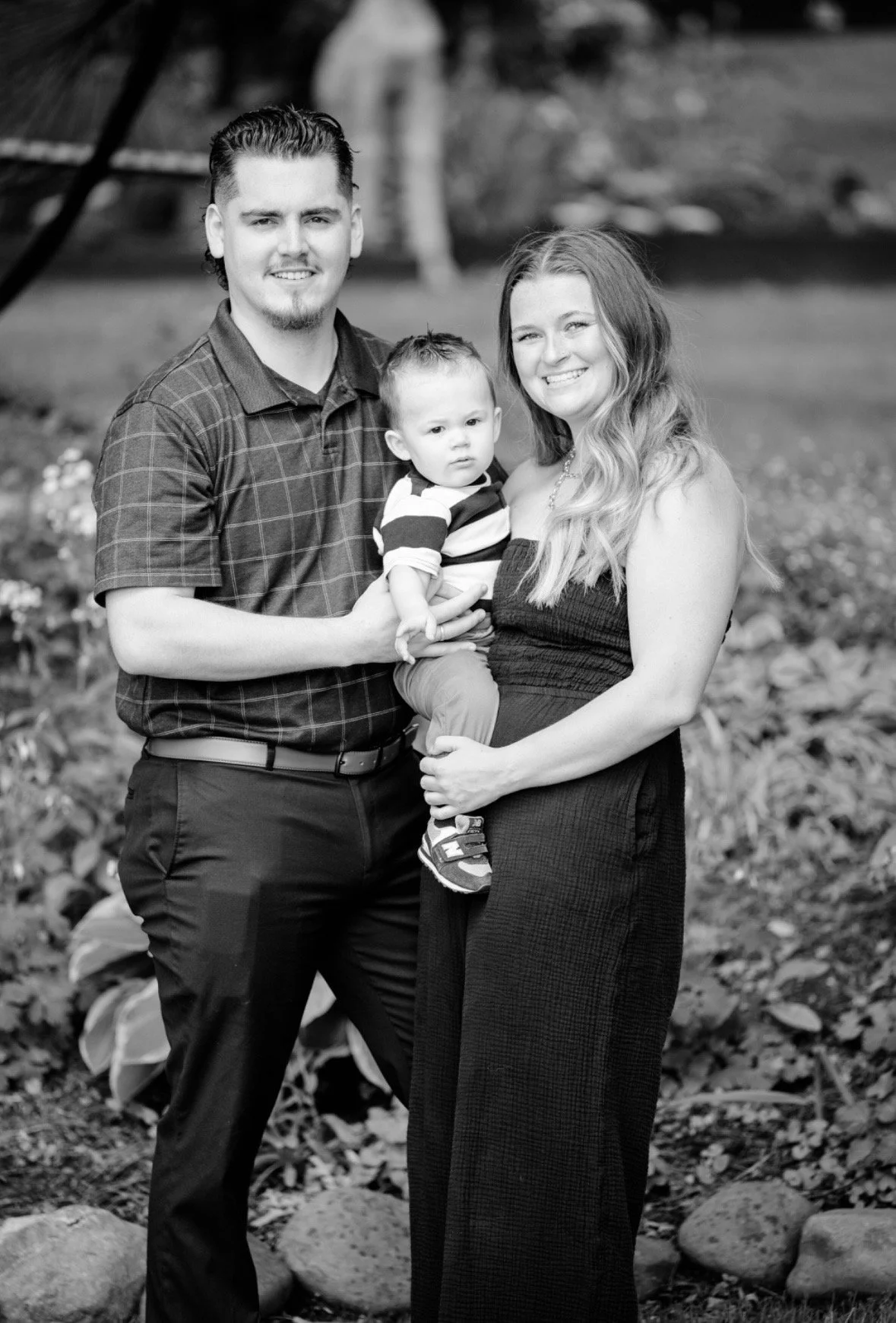 Black and white photo of a family of three outdoors, with a man, a woman, and a young child. The man has short, styled hair and is wearing a checkered shirt. The woman has long wavy hair and is wearing a strapless dress. The child, being held by the woman, has short hair and is wearing a striped shirt and pants.
