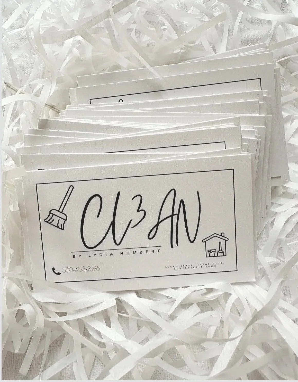 A stack of business cards with the name 'CLEAN' by Lydia Humbert, featuring icons of a broom and cleaning supplies, placed in shredded white paper.