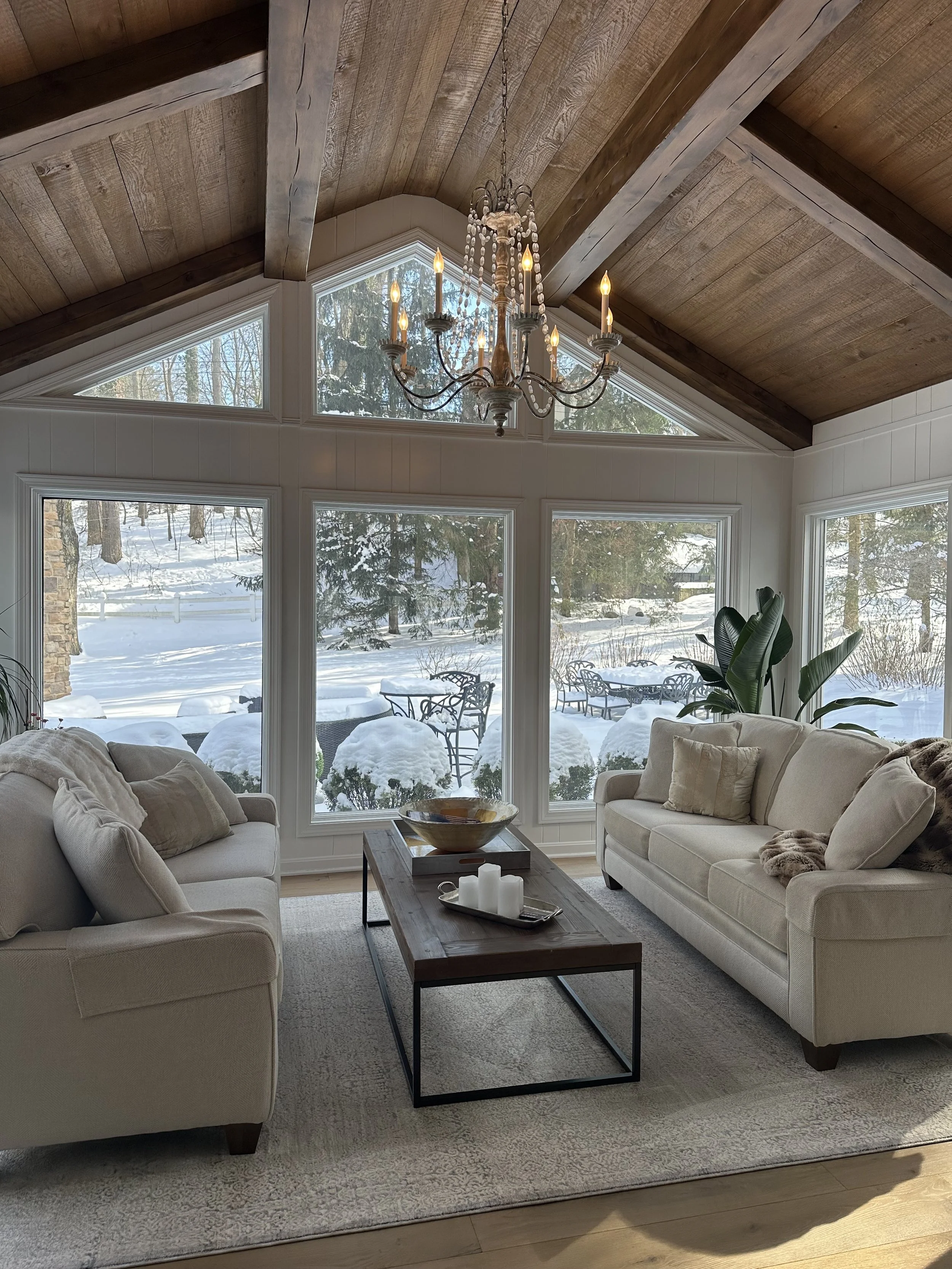 Living room with large windows showing a snowy outdoor scene, two beige sofas, a wooden coffee table, a chandelier, a large potted plant, and cozy pillows and throws.