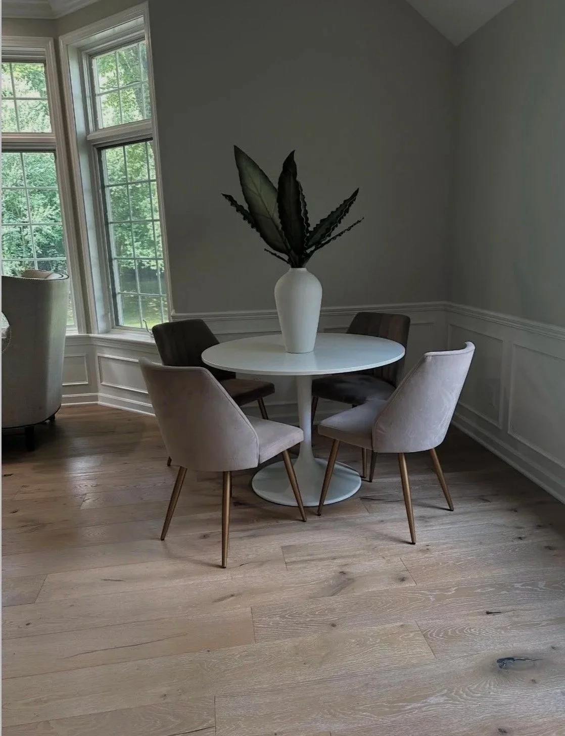 A dining area with a round white table, three beige chairs, and a white vase with large green leaves on the table. Windows showing greenery outside.
