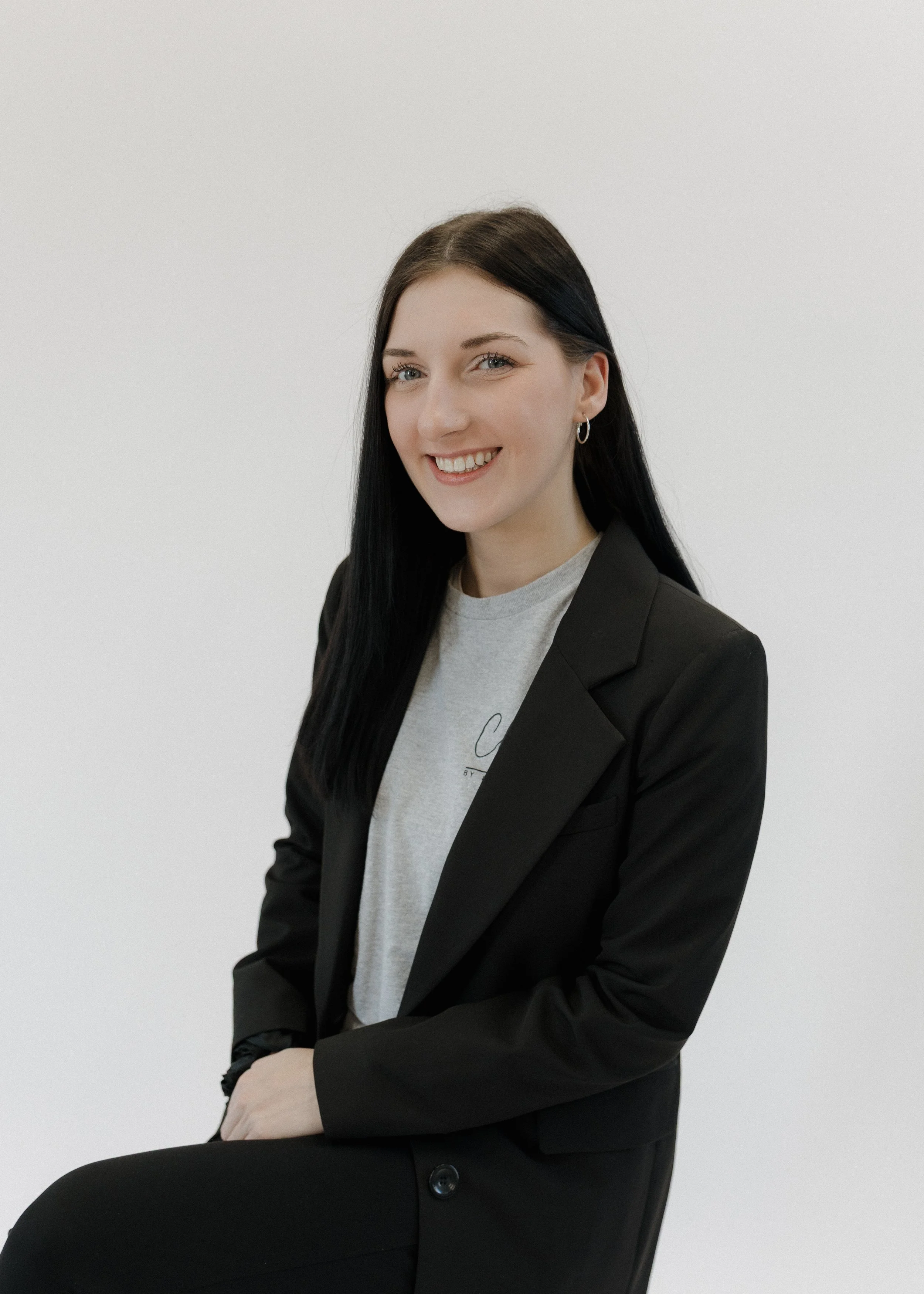 A young woman with long black hair, smiling, wearing a black blazer over a light gray t-shirt, sitting against a plain white background.