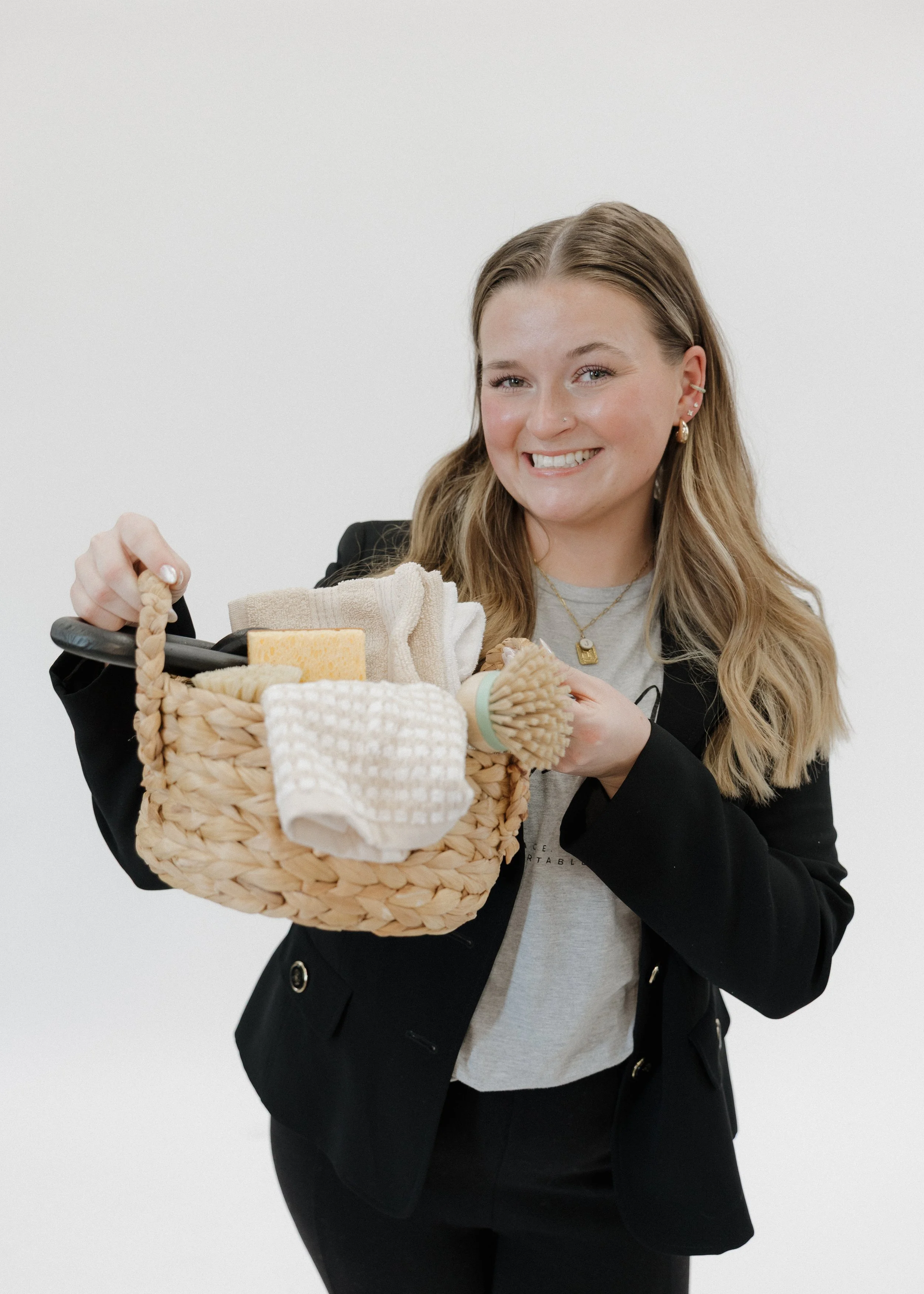 A young woman with long blonde hair smiling, holding a woven basket filled with various household cleaning items and textiles, against a plain white background.