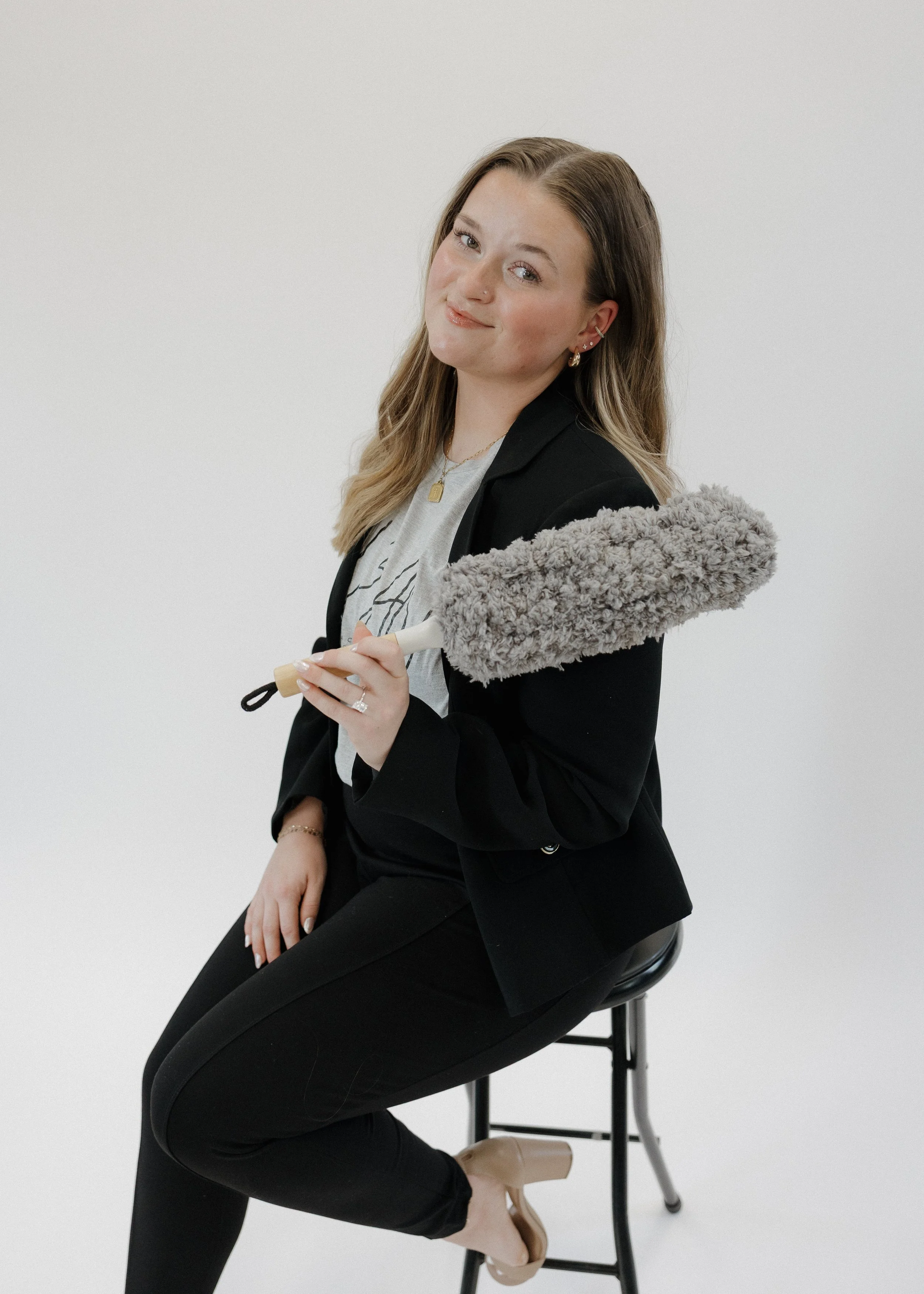 A woman with light brown hair, wearing a black blazer and black pants, sitting on a black stool with a white background. She is holding a duster with a fluffy gray head and smiling at the camera.