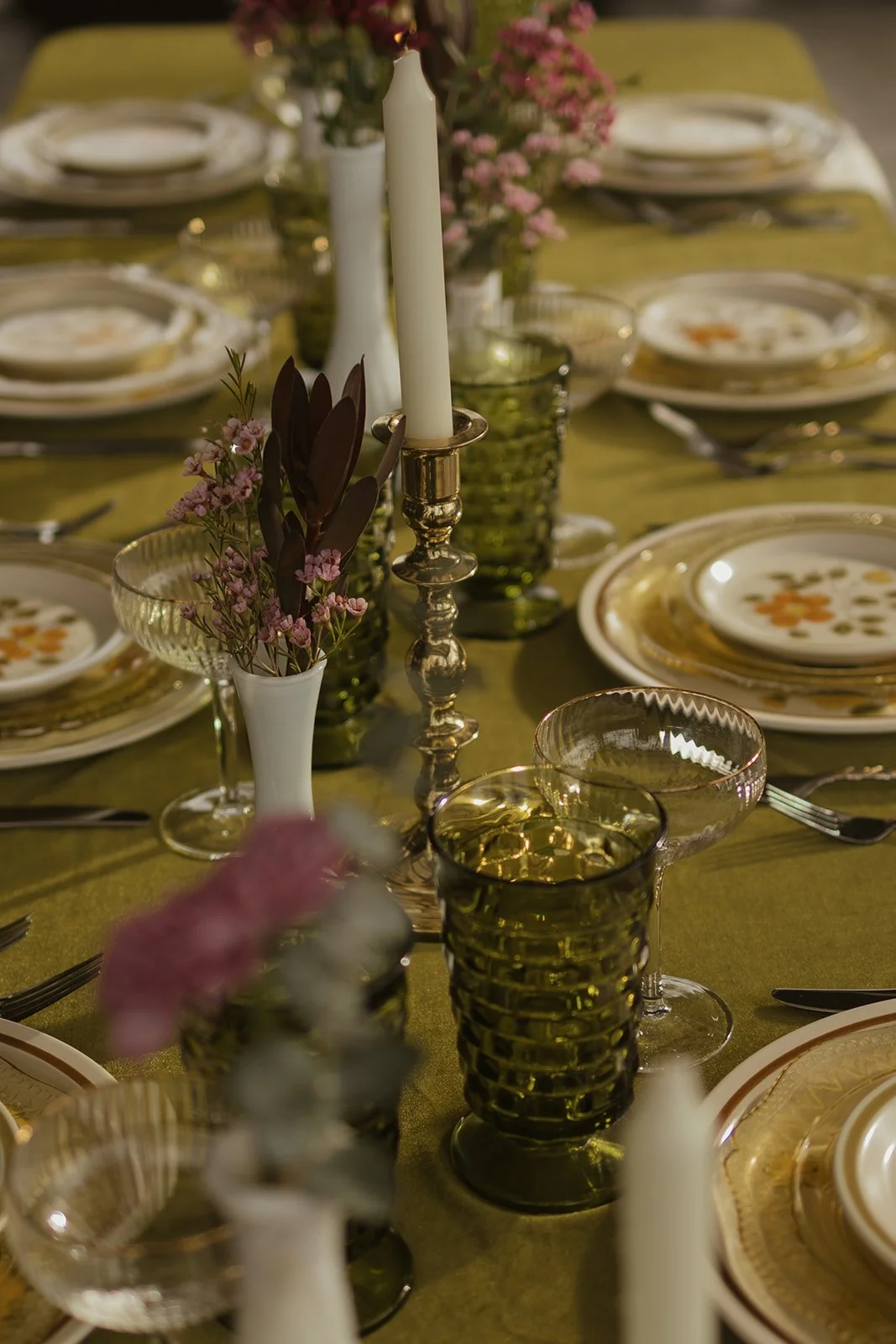 A decorated dining table with green tablecloth, stoneware plates, glassware, pink and purple flowers in vases, and lit white candles.
