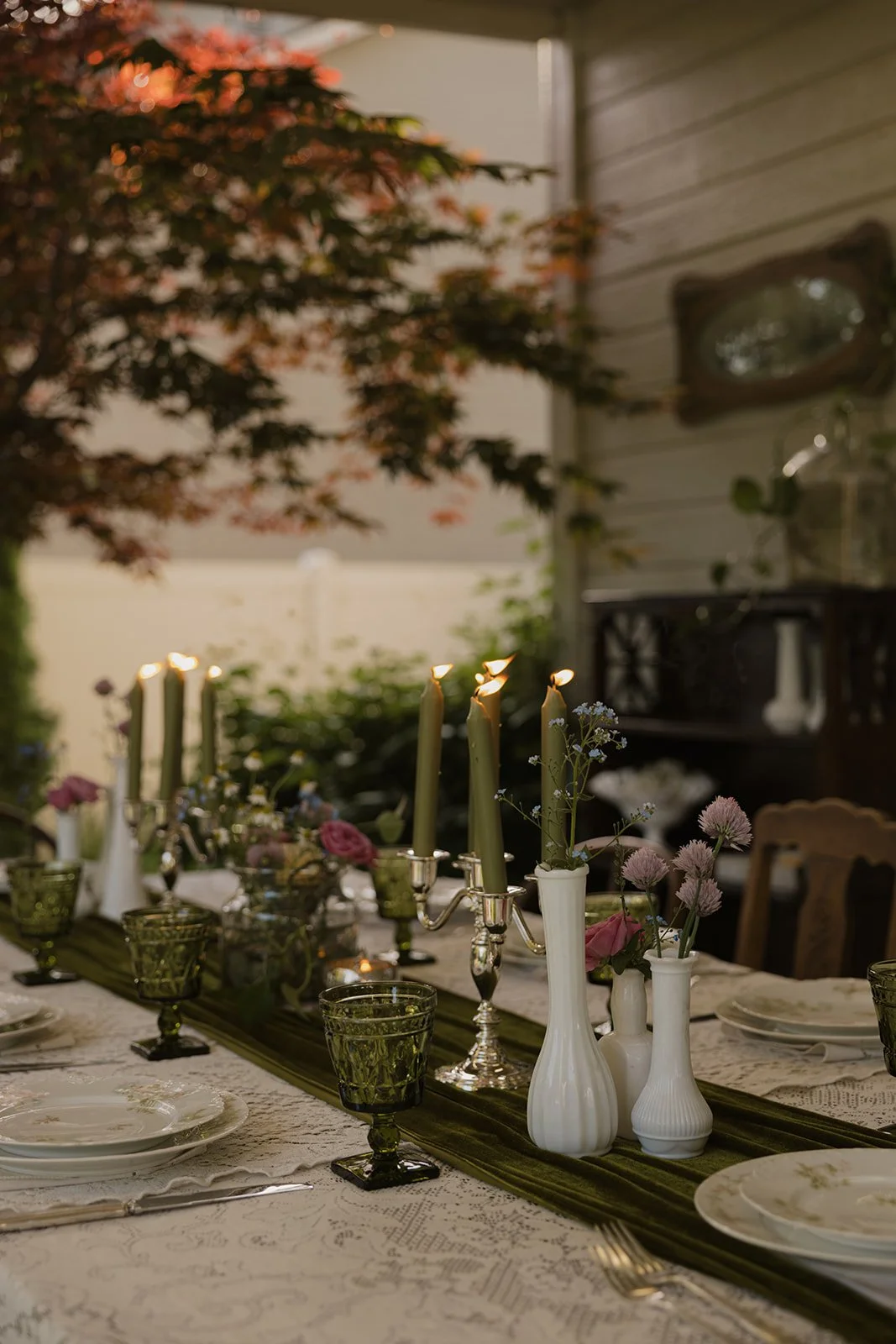 A dinner table set for a gathering outdoors with candles, flowers, and vintage green glasses, surrounded by greenery.