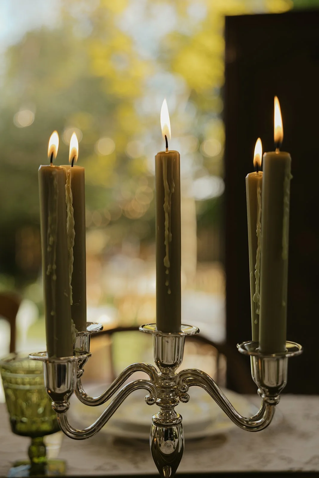 A silver candelabrum with five lit candles, melted wax dripping down, set on a table with a blurred outdoor background.