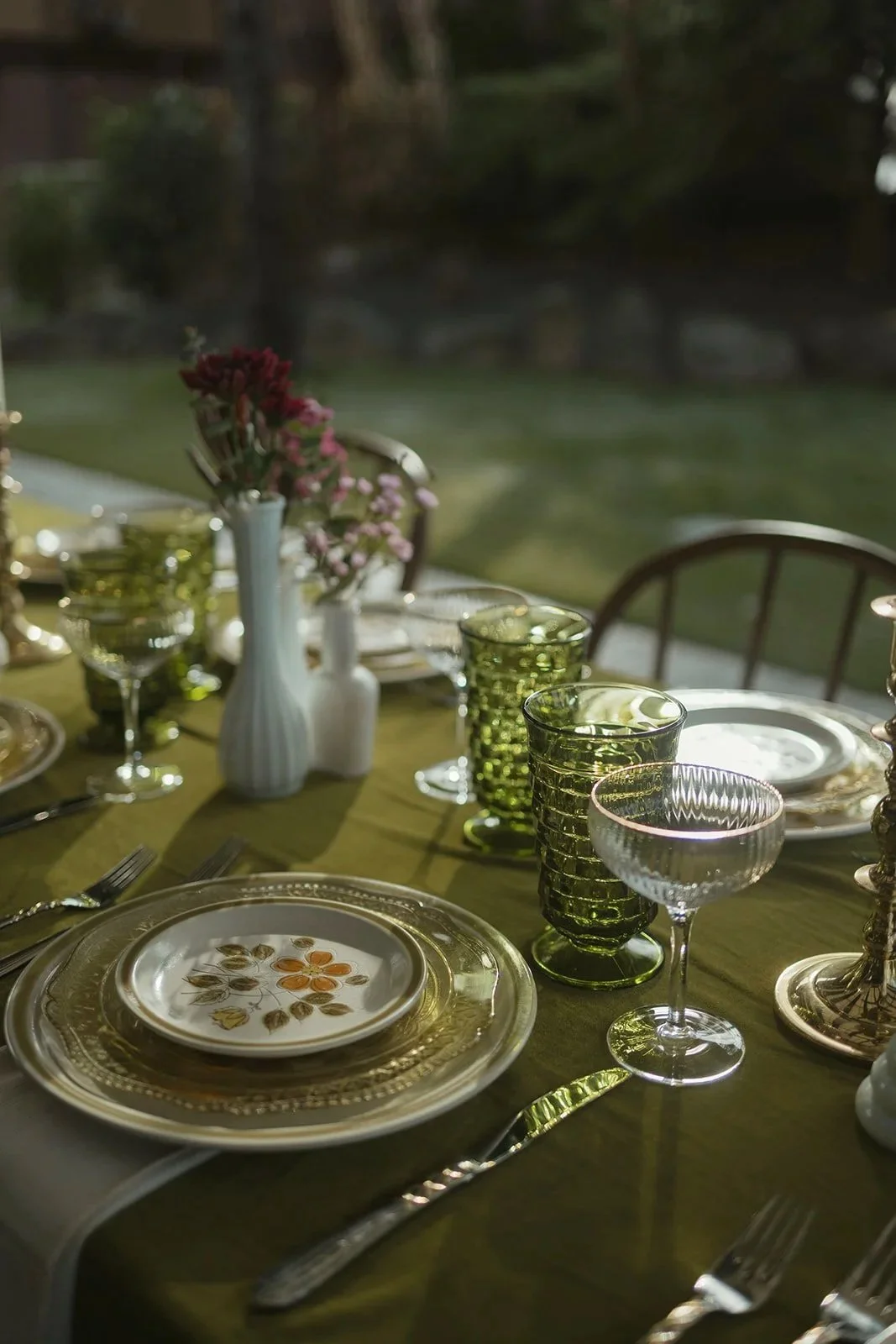 Elegant outdoor dining table set with vintage plates, green textured glassware, silver cutlery, a white vase with pink and red flowers, and a backdrop of a garden with fading daylight.