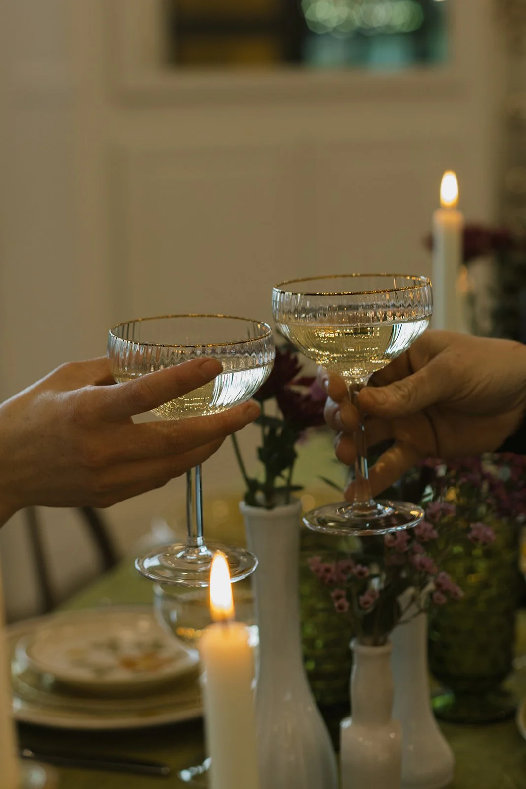 Two people clinking glasses of white wine during a dinner celebration with lit candles and floral arrangements in the background.