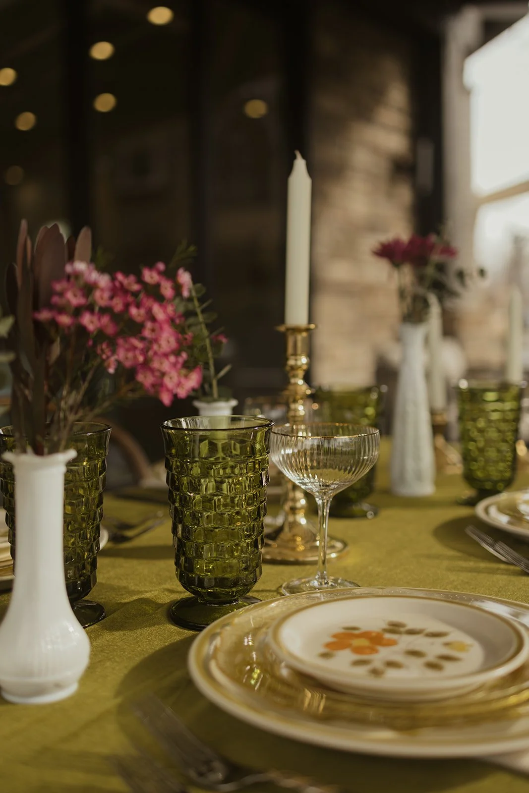 A dining table set with green textured glasses, clear glassware, white candles, pink flowers in vases, and decorative plates on a green tablecloth.