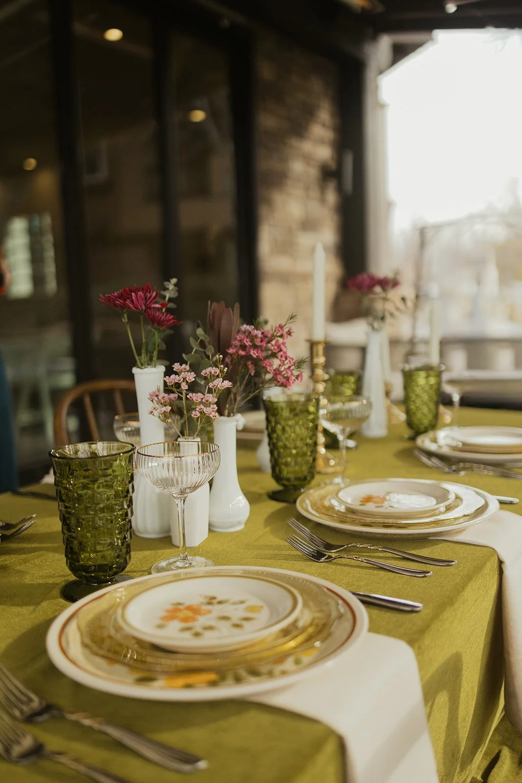 A retro tablescape, set with floral arrangements, green glassware, and stoneware plates, featuring an outdoor setting with sunlight streaming through a window.