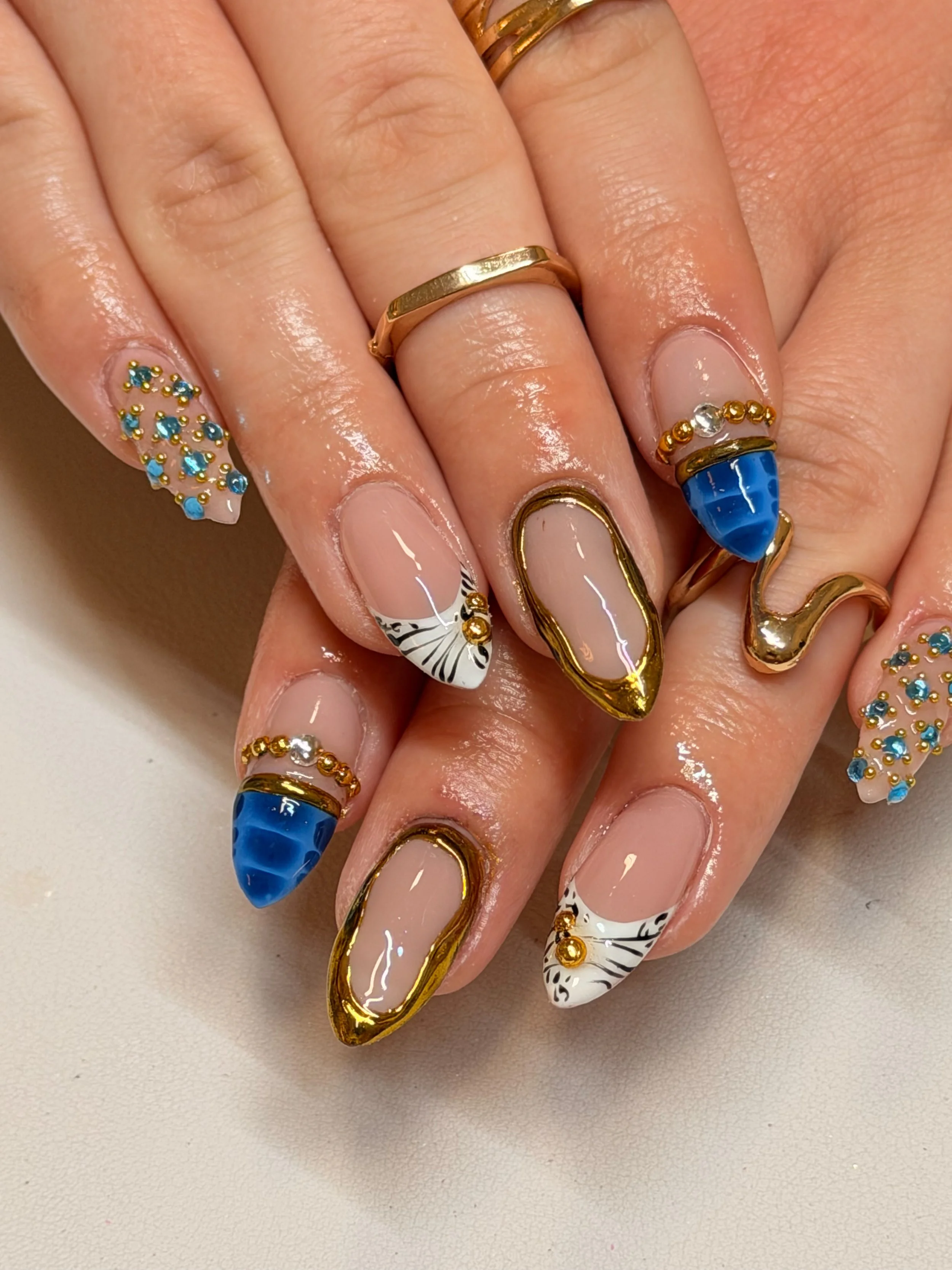 Close-up of hands with elaborate nail art featuring gold accents, blue stones, rhinestones, and black and white designs, adorned with gold rings.