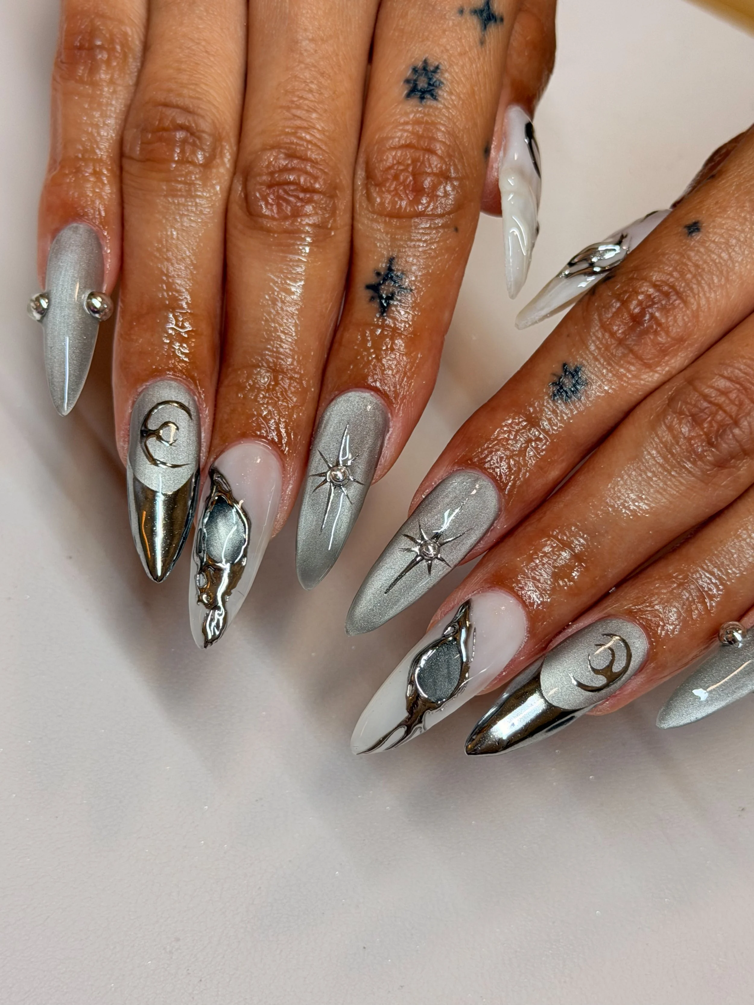 Close-up of manicured nails with metallic silver, white, and black nail polish, decorated with stars, moons, and metallic accents.