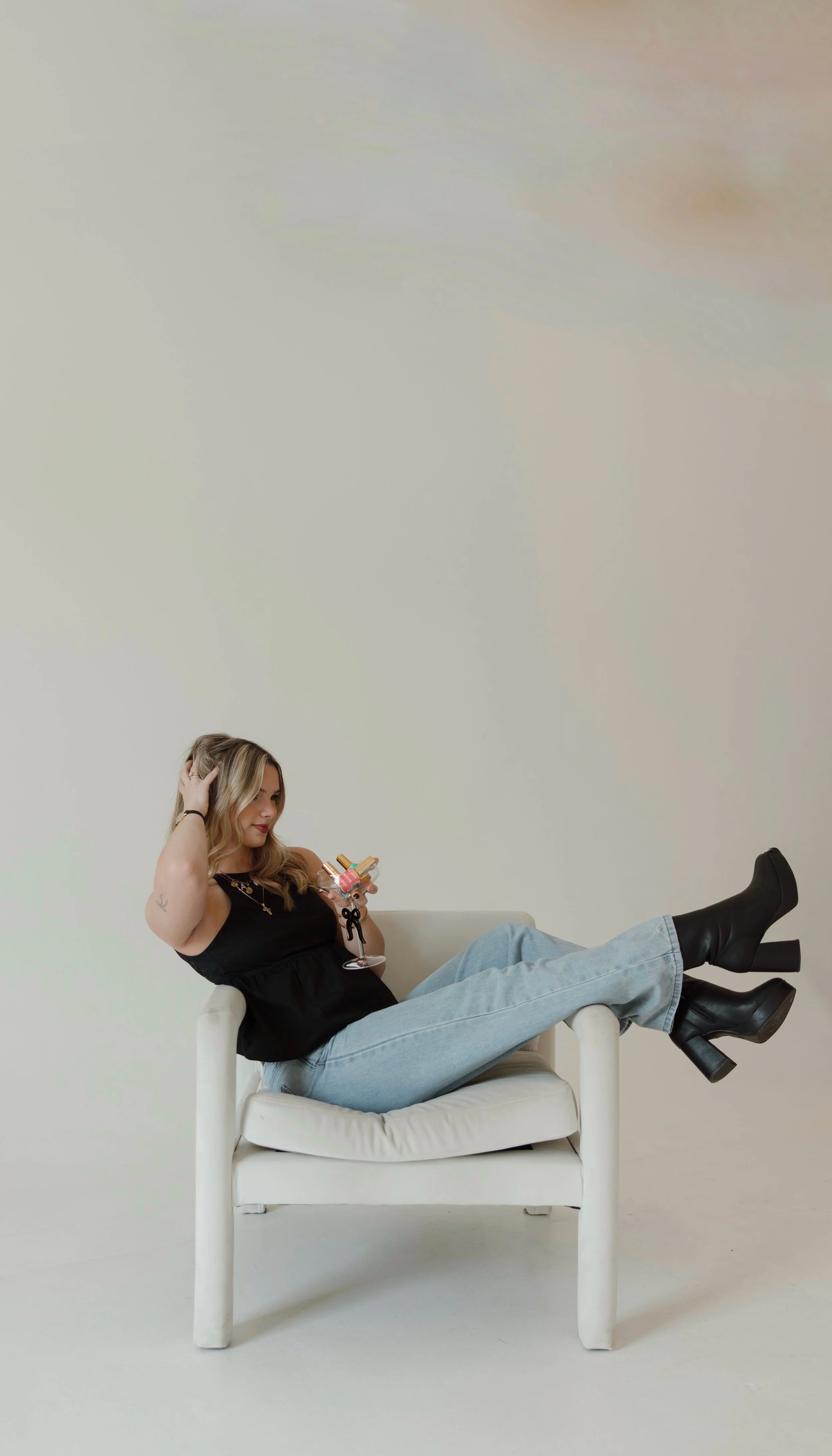 Woman with blonde hair sitting on a white chair, wearing a black top, light blue jeans, and black heeled boots, holding a glass of pink drinks, with one hand in her hair, against a plain light background.