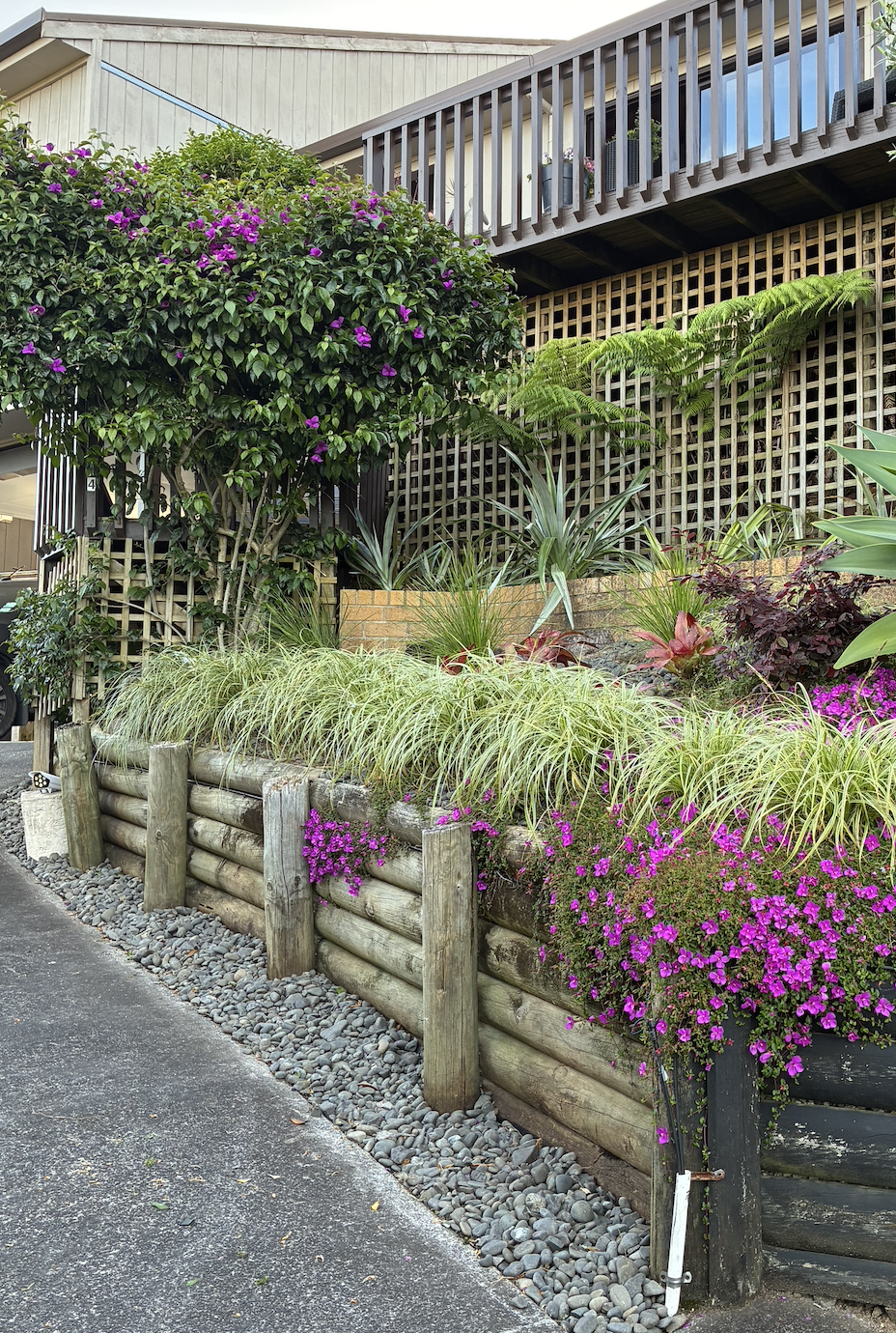 A landscaped driveway raised garden with a variety of easycare plants and flowers, bordered by a wooden retaining wall with a newly weeded out base with new weedmatting pinned in place and topped with claened existing riverstones.