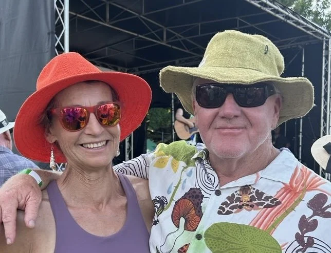 A smiling woman and man posing outdoors, both wearing colorful hats and sunglasses. The woman has a red hat and pink mirrored sunglasses, and the man has a green hat and black sunglasses. They are standing close together, with the woman’s arm around the man.