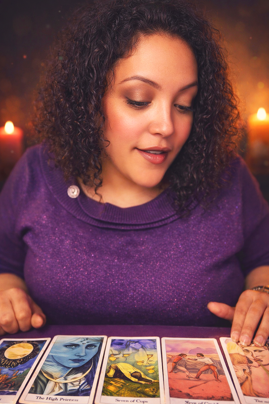 A woman with curly hair and a purple shirt looks at tarot cards laid out on a table. There are candles in the background.