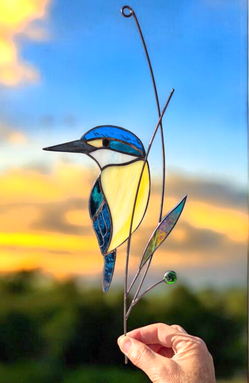 Stained glass artwork of a kingfisher bird perched on a branch with a sunset sky in the background.