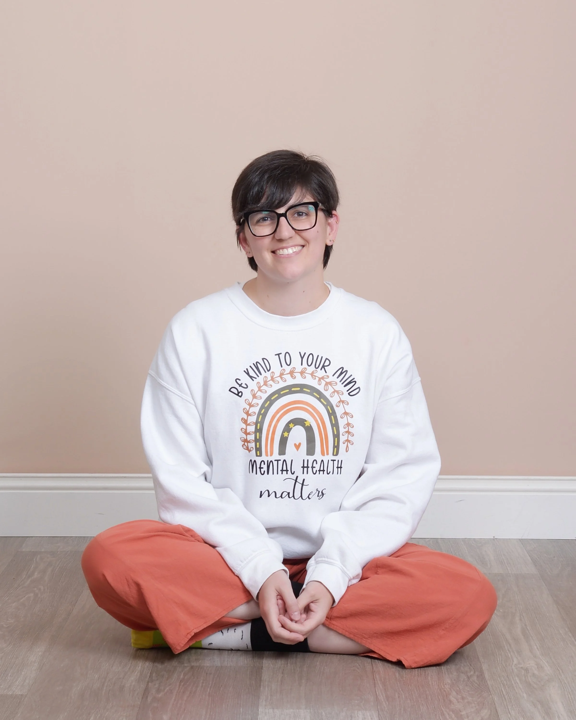 Kirsten Dantin LPC Headshot. Person sitting cross-legged on the floor, wearing glasses, a white sweatshirt with a colorful rainbow and text about mental health, and orange pants. They are smiling and sitting against a beige wall.