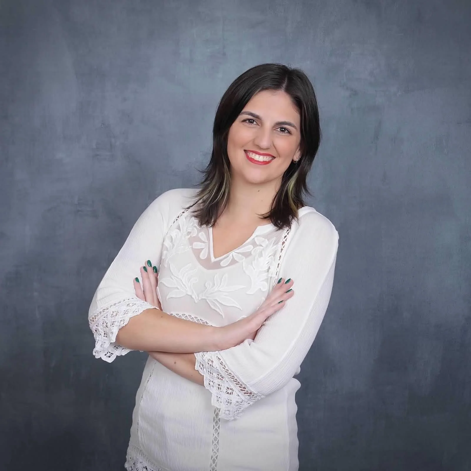 Kirsten Dantin LPC Headshot. A woman with shoulder-length dark hair, smiling, wearing a white dress with lace details and crossed arms, standing against a gray textured background.