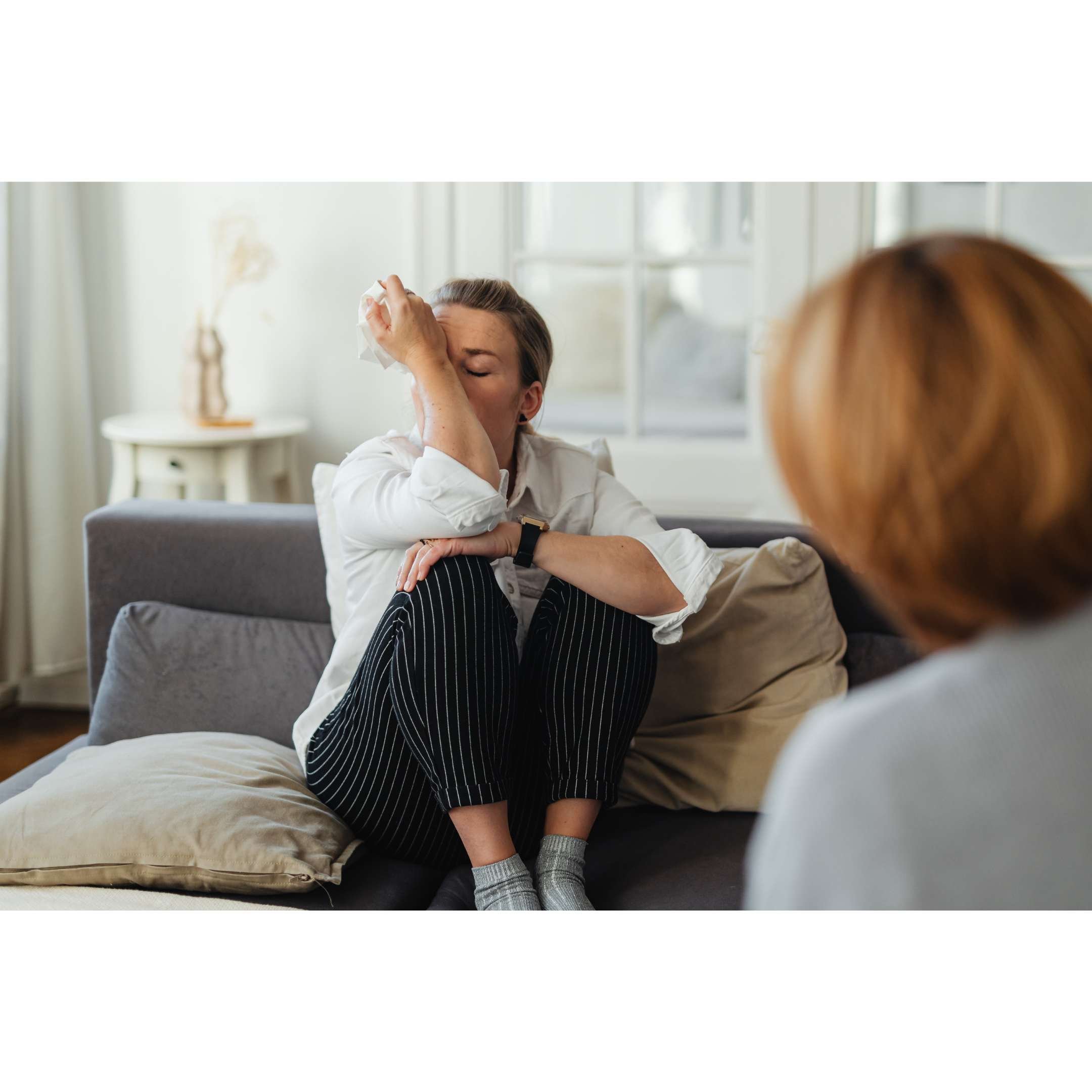 A woman sitting on a couch, crying and wiping her eyes with a tissue, while another woman listens attentively.
