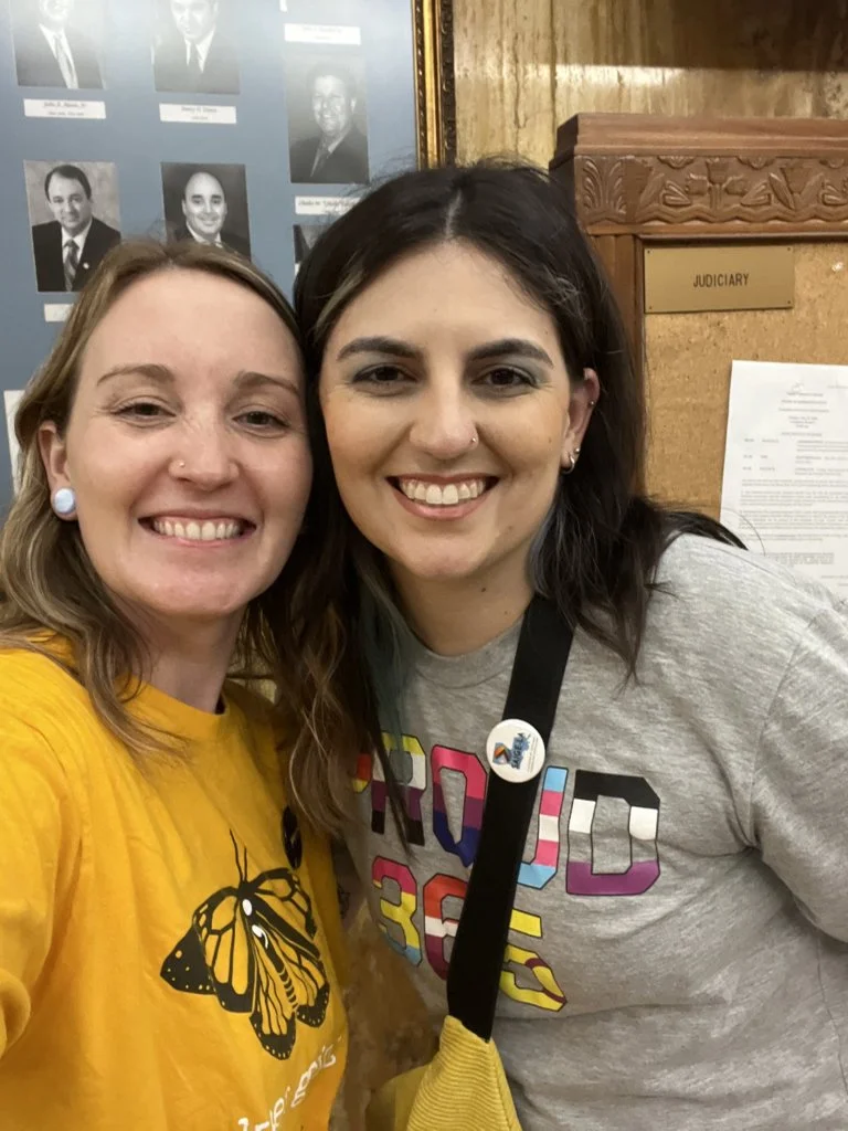 Two women smiling, taking a selfie indoors near a wooden jury box with a sign labeled 'JUDICIARY' behind them. One woman wears a yellow shirt with a butterfly design, and the other wears a gray shirt with colorful block letters. There is a bulletin board with papers and a framed photo collage in the background.