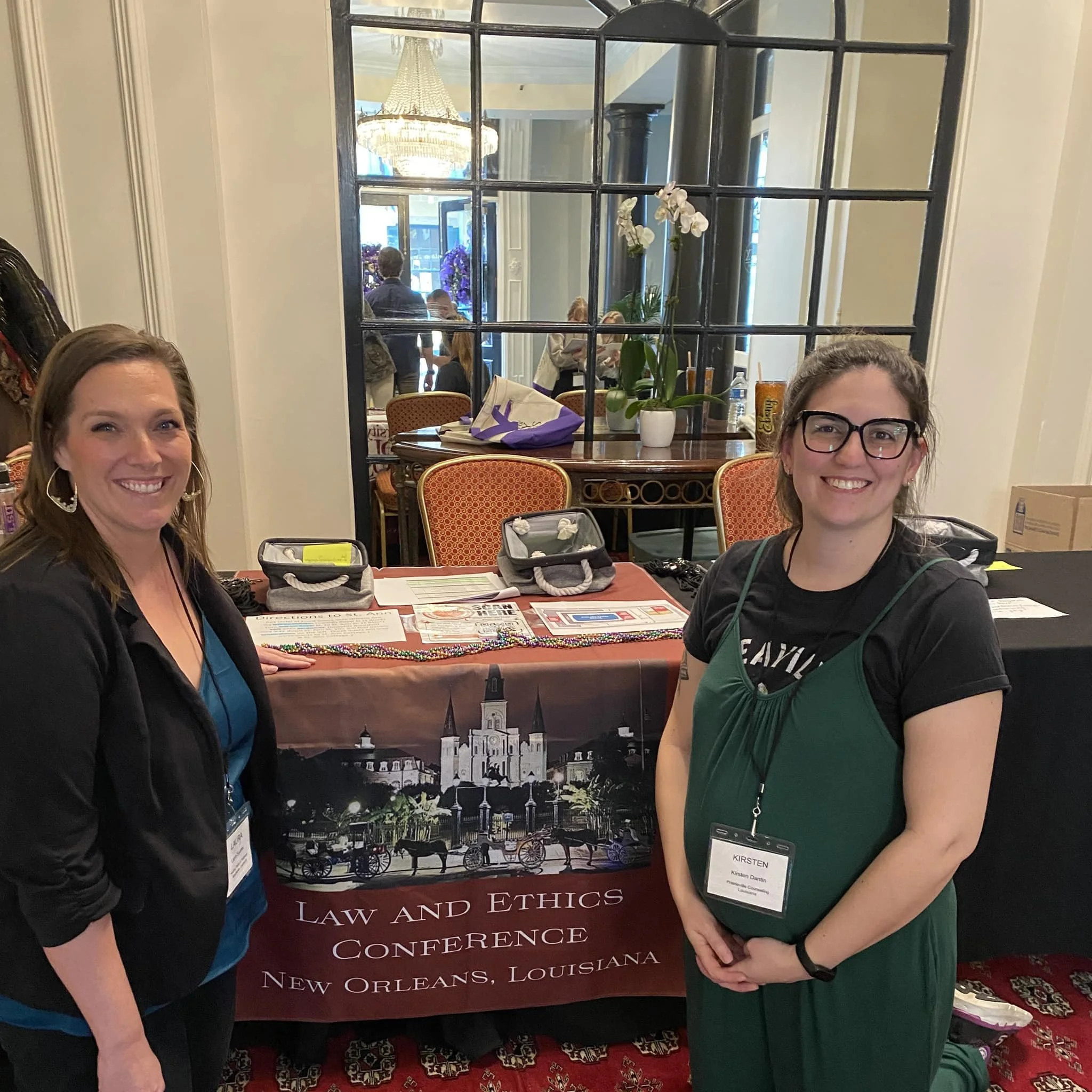 Two women standing in front of a table at a law and ethics conference in New Orleans, Louisiana. The table has brochures and a Mardi Gras bead necklace. A large mirror, chandelier, and some people are visible in the background.