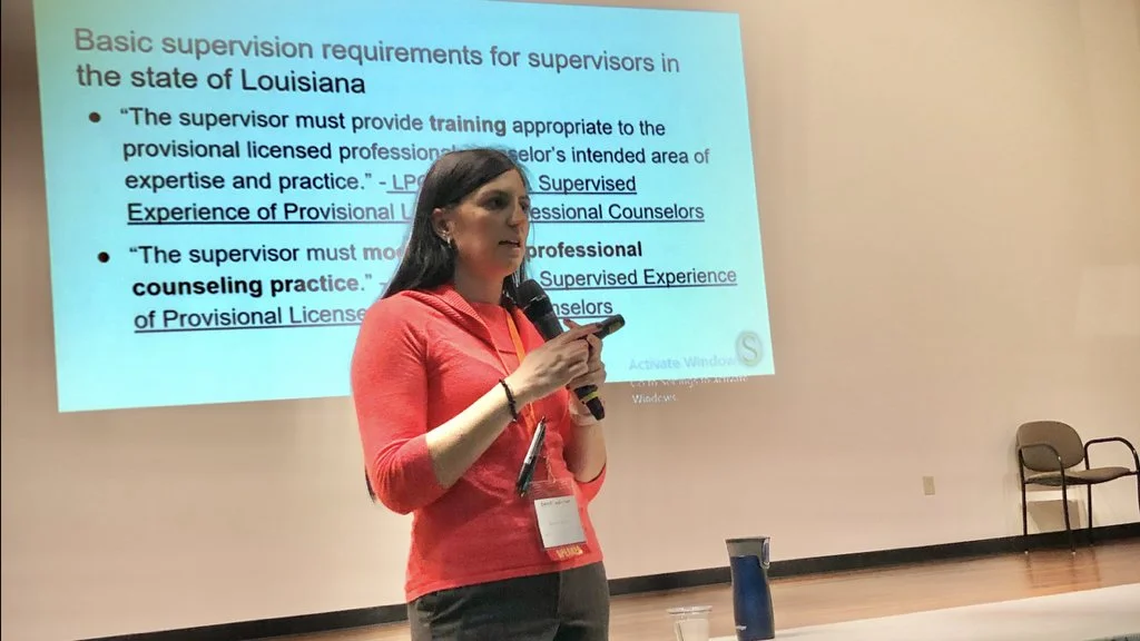 A woman with dark hair, wearing a red top and holding a microphone, presenting a slide on supervision requirements for counselors in Louisiana, projected on a screen in a conference room.
