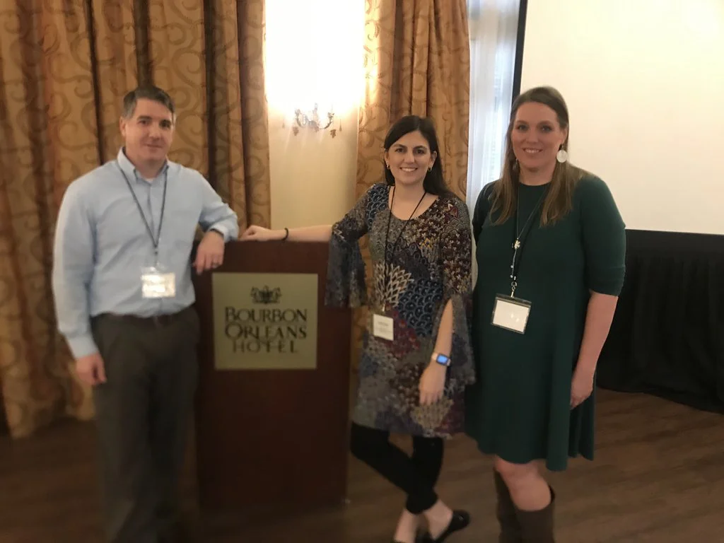 Three people standing inside a hotel ballroom, with a sign for Bourbon Orleans Hotel, smiling at the camera.