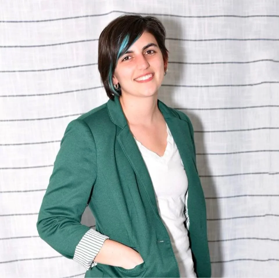 Kirsten Dantin LPC Headshot. A woman with short dark hair and a streak of turquoise, wearing a green blazer and a white top, smiling at the camera, standing in front of a striped curtain.