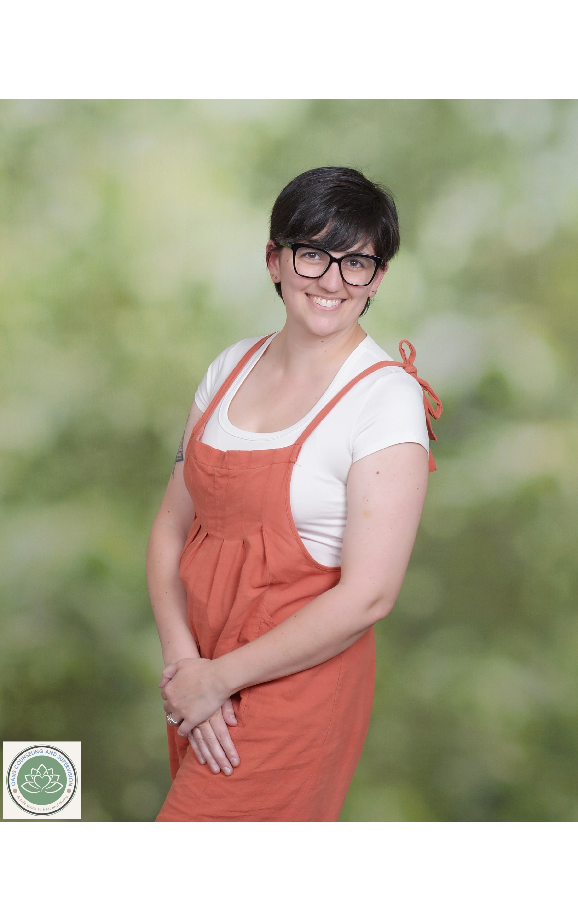 Kirsten Dantin LPC Headshot. A smiling woman with short dark hair, glasses, wearing a white t-shirt and an orange apron, stands against a blurred green nature background.