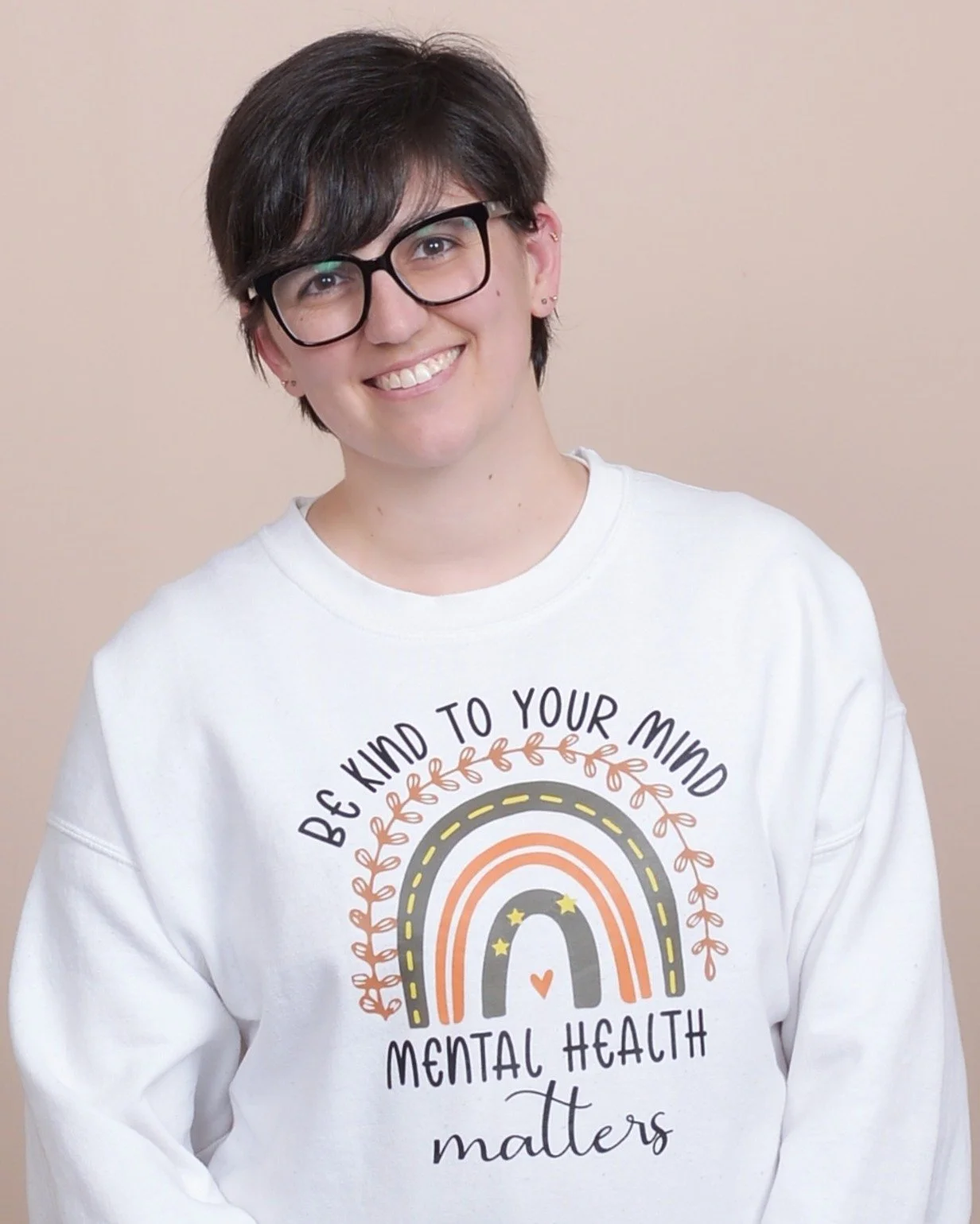 Kirsten Dantin LPC Headshot. A person with short dark hair and glasses smiling, wearing a white sweatshirt with a rainbow design and the phrase 'Be kind to your mind, mental health matters' printed on it, standing against a beige background.
