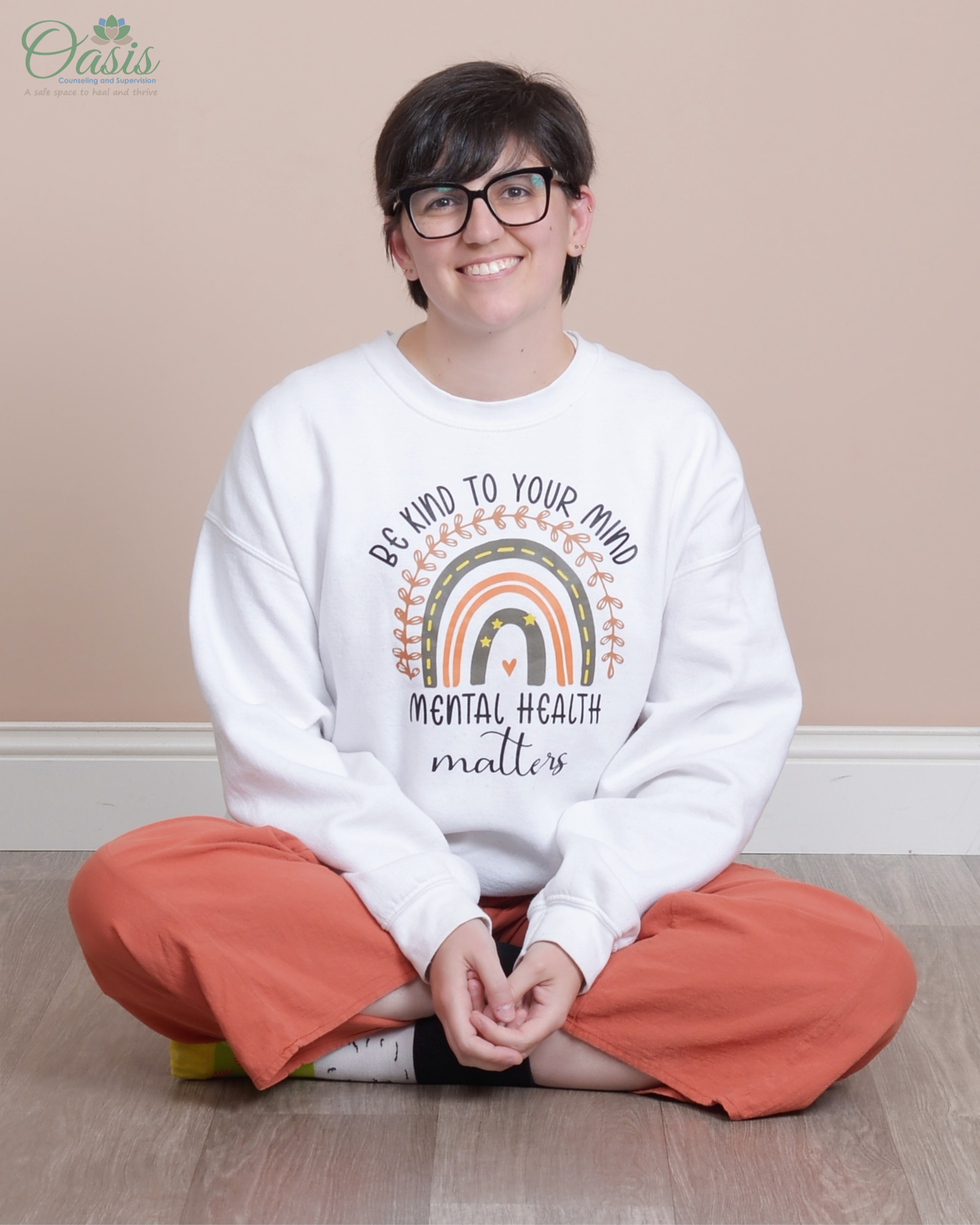 Kirsten Dantin LPC Headshot. She is sitting cross-legged on a wooden floor, wearing a white sweatshirt with text and design about mental health, and orange pants, with a logo saying 'Oasis Counseling and Supervision' in the top left corner.