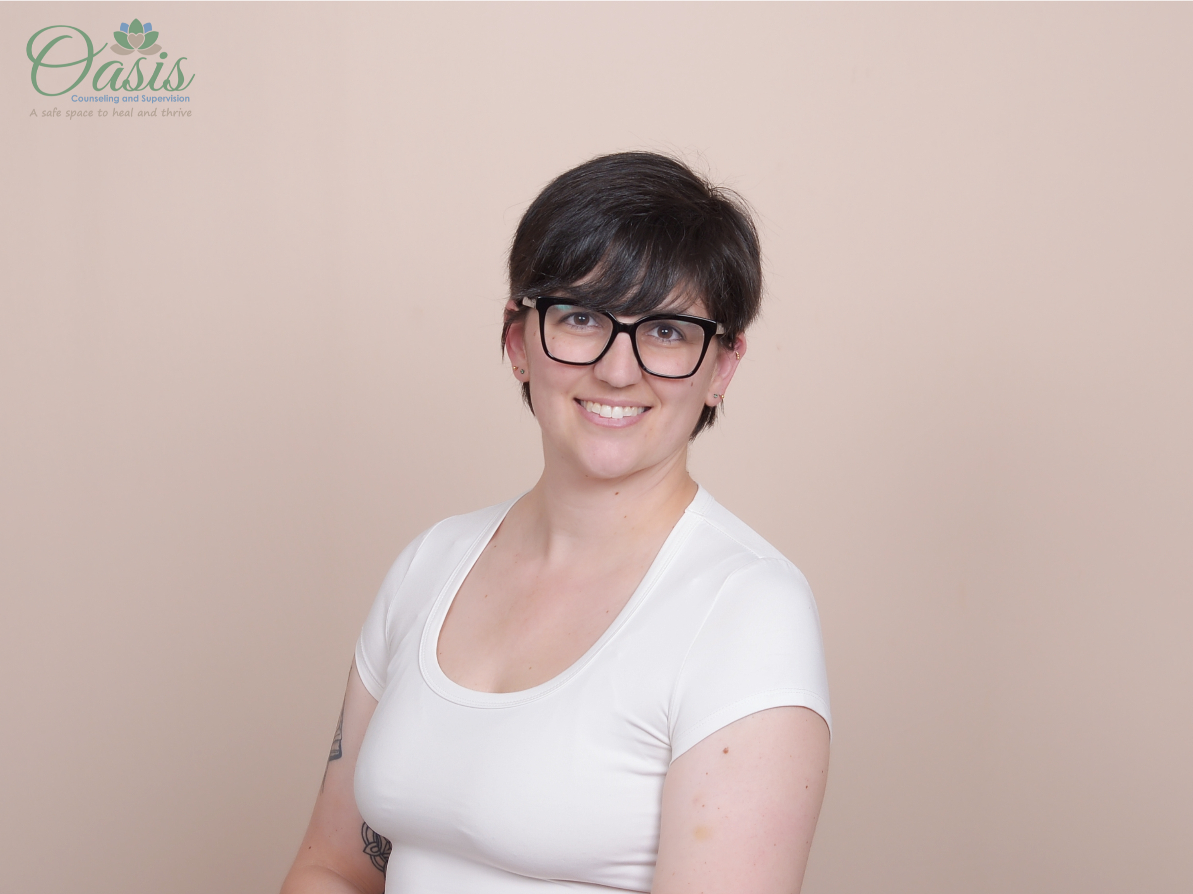Kirsten Dantin LPC Headshot. She is a young woman with short dark hair, glasses, and earrings, smiling, wearing a white top, standing against a beige background with "Oasis Counseling and Supervision" logo in the top left corner.