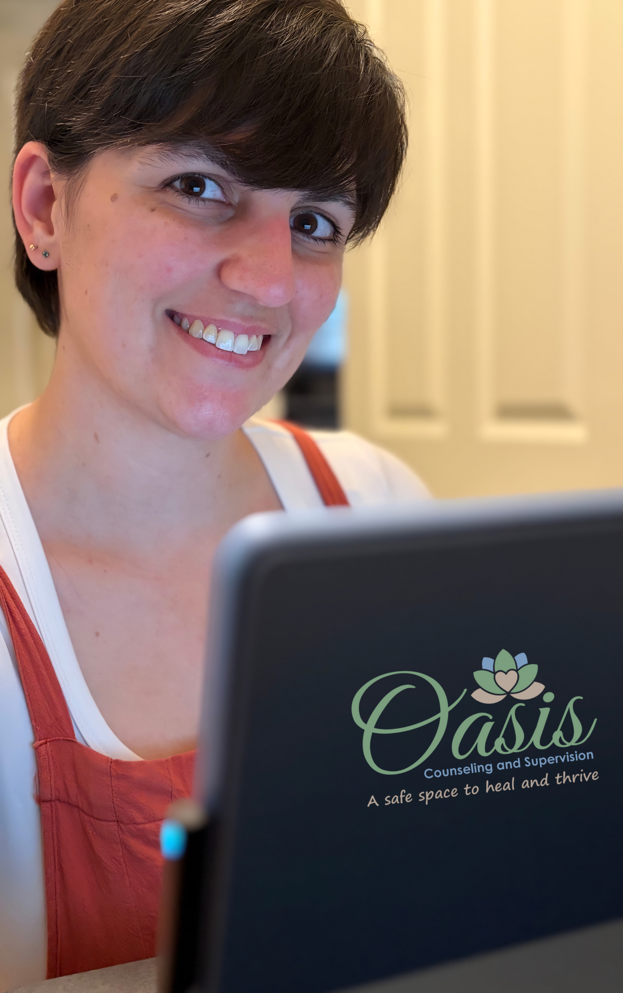 A smiling woman looking at a laptop screen displaying 'Oasis Counseling and Supervision' with a lotus flower logo on a blurred background.