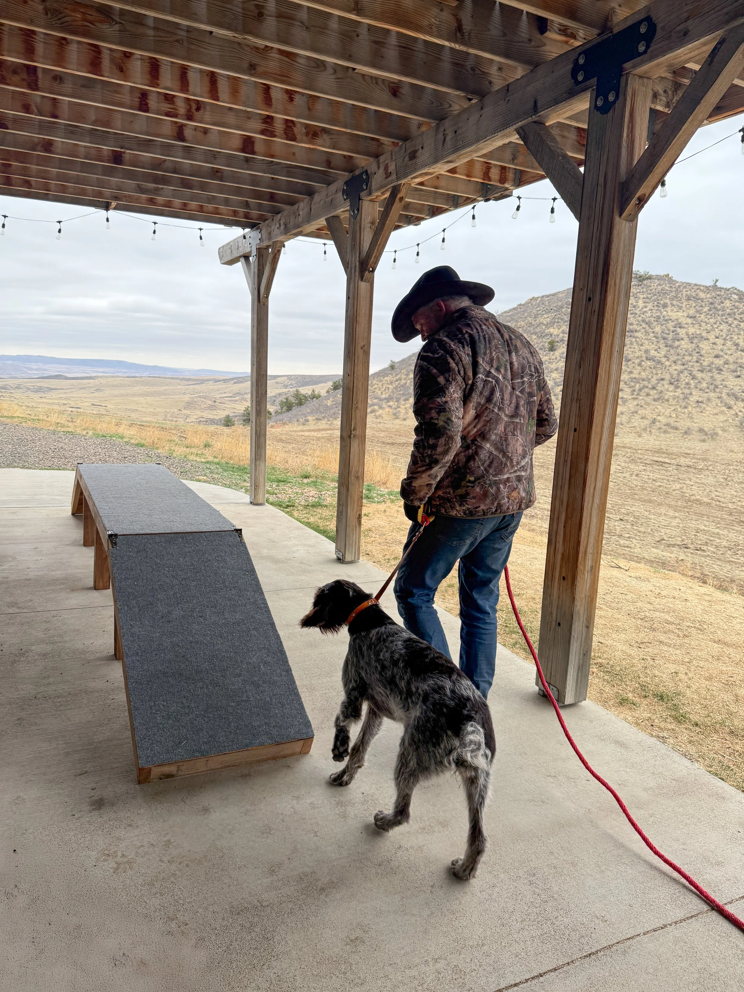 A man in a cowboy hat and camouflage jacket with a black and white dog on a leash under a wooden shelter, overlooking a countryside landscape with hills and open fields.