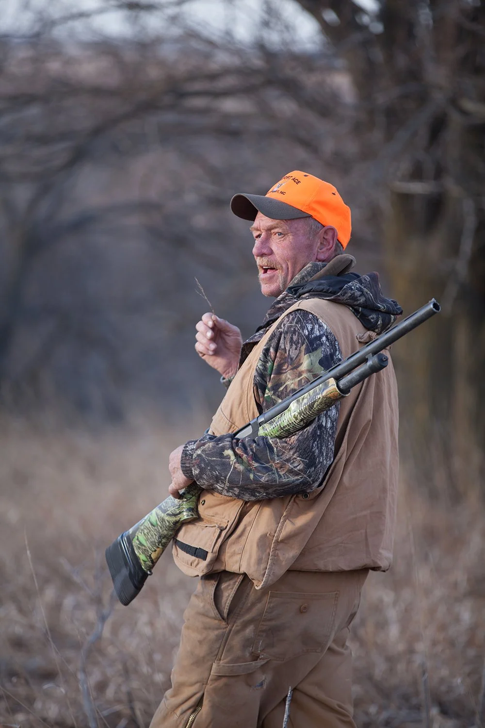 A man in camouflage and tan outdoor gear, wearing an orange cap, holding a hunting rifle over his shoulder, standing outdoors in a wooded area.